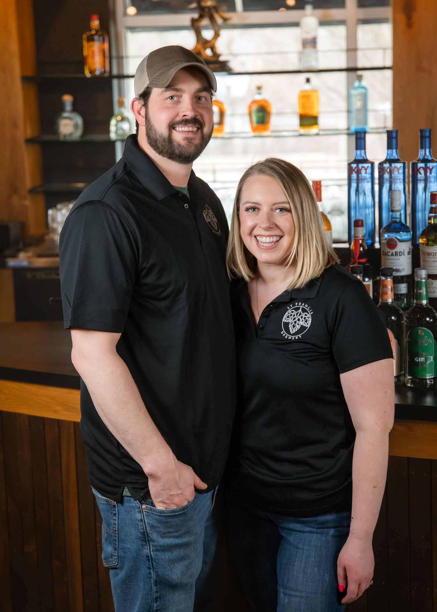 Two staff members in black shirts and jeans pose together in front of a bar counter with liquor bottles visible.