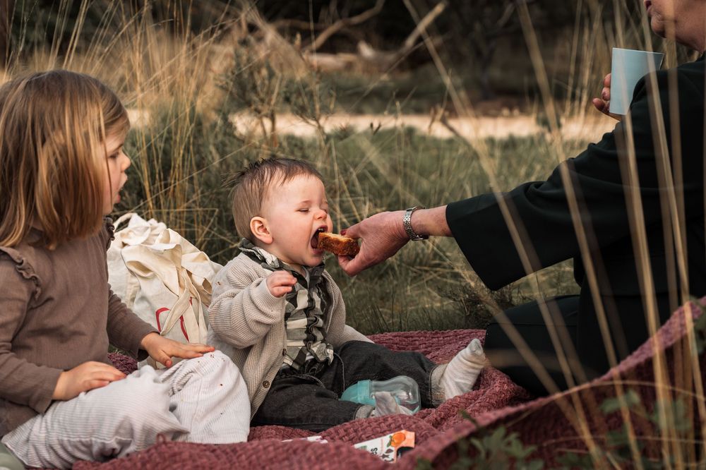 A person in a brown sweater holds a small child outdoors during autumn while surrounded by tall grass and trees.