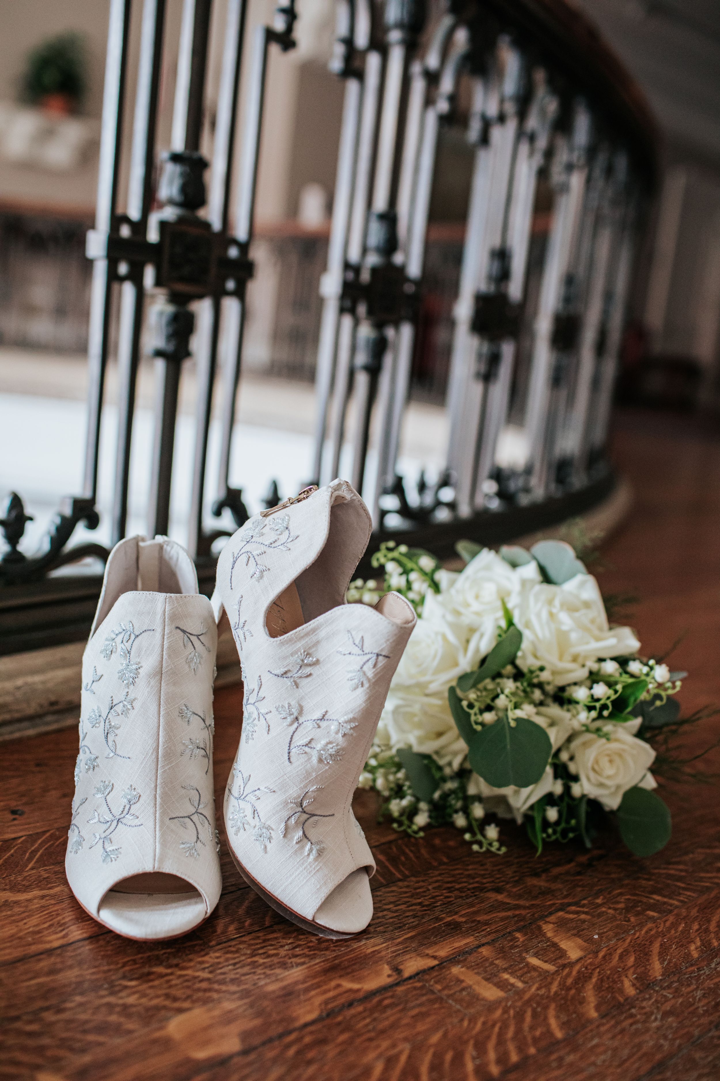 White lace bridal booties with peep toes sit beside an elegant white rose and eucalyptus bouquet on a wooden floor.