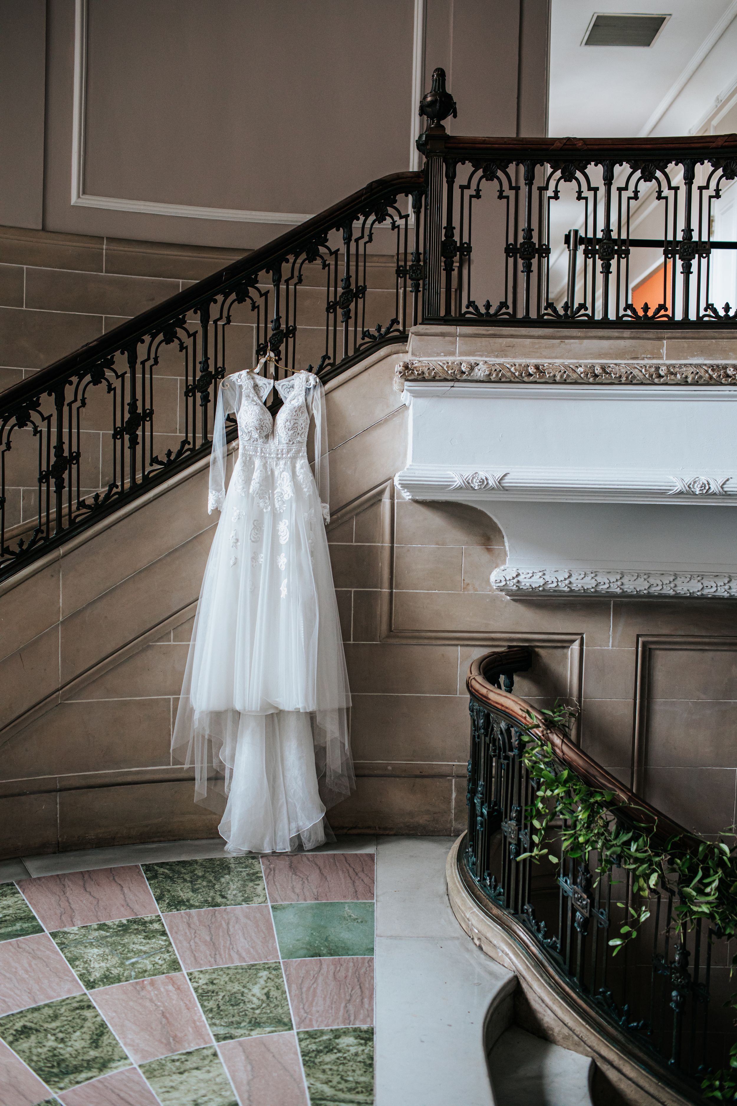 White wedding dress displayed on ornate staircase with wrought iron railing and marble floors in elegant venue.
