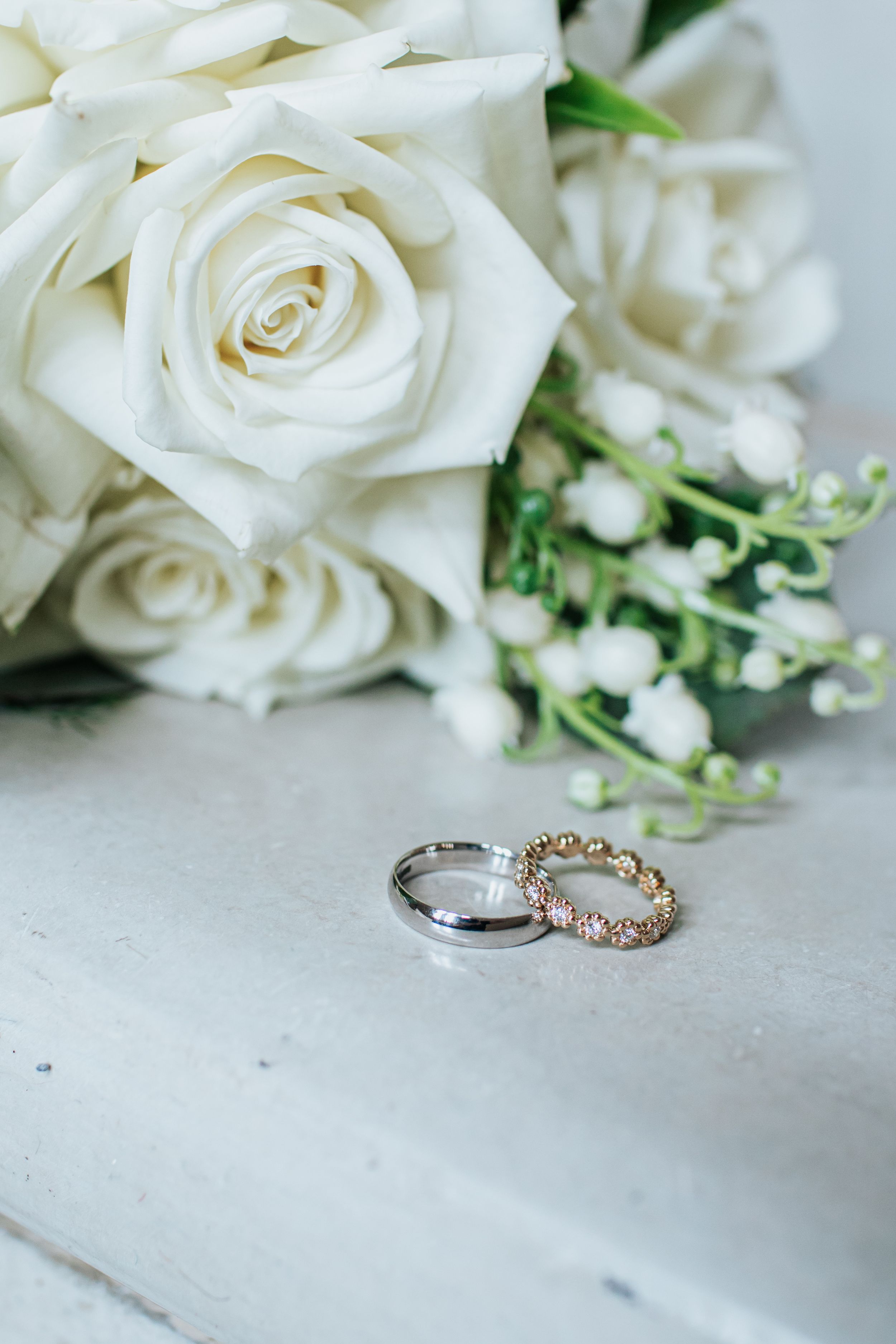 White roses and delicate wedding rings displayed on a light marble surface in an elegant bridal arrangement.