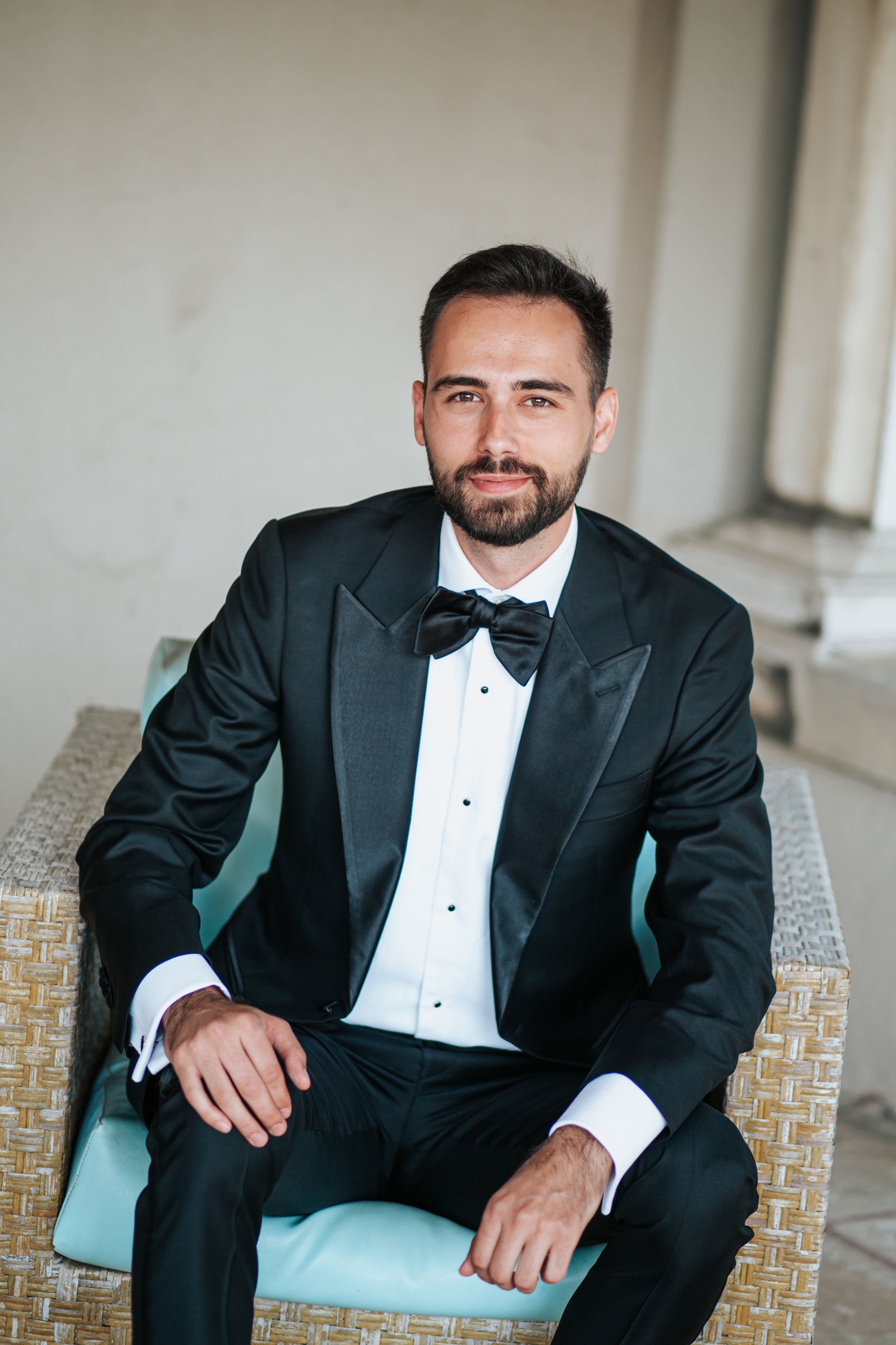 A groom in a classic black tuxedo and bow tie sits on a light blue chair during formal wedding portraits.
