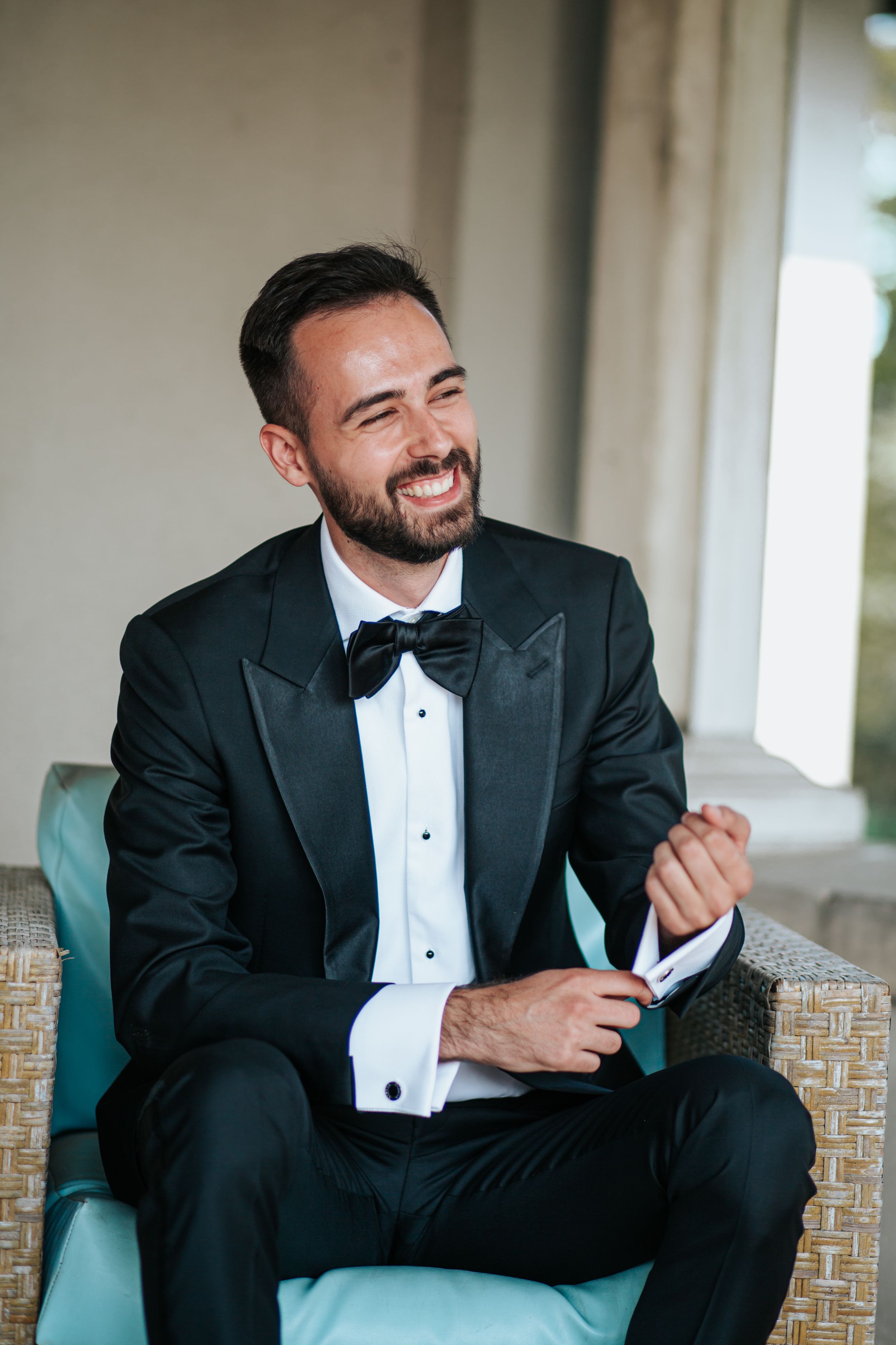 Smiling groom in classic black tuxedo and bow tie sits on turquoise chair during formal wedding portraits.