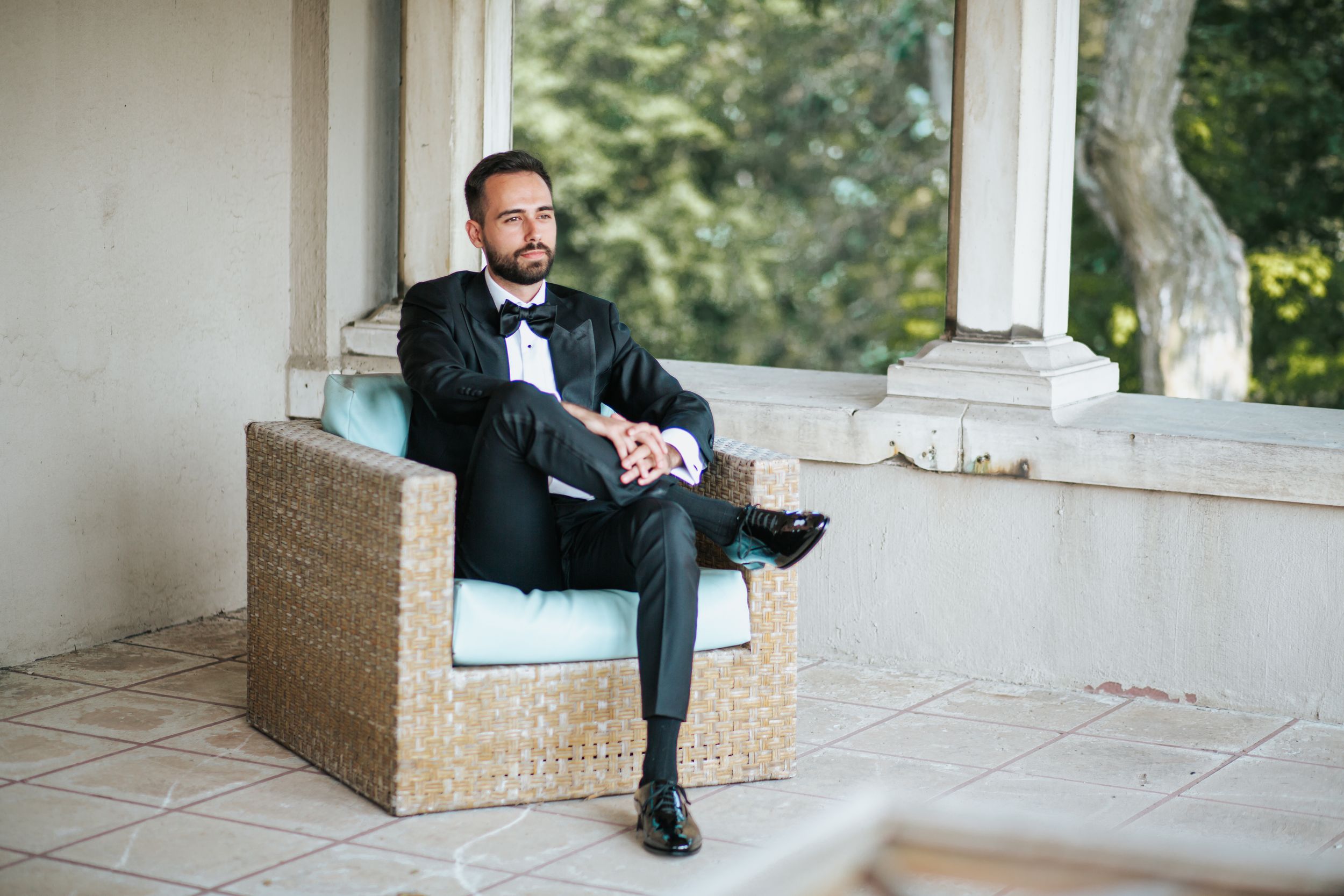 A person in a black tuxedo sits casually on a wicker outdoor sofa on a covered patio.