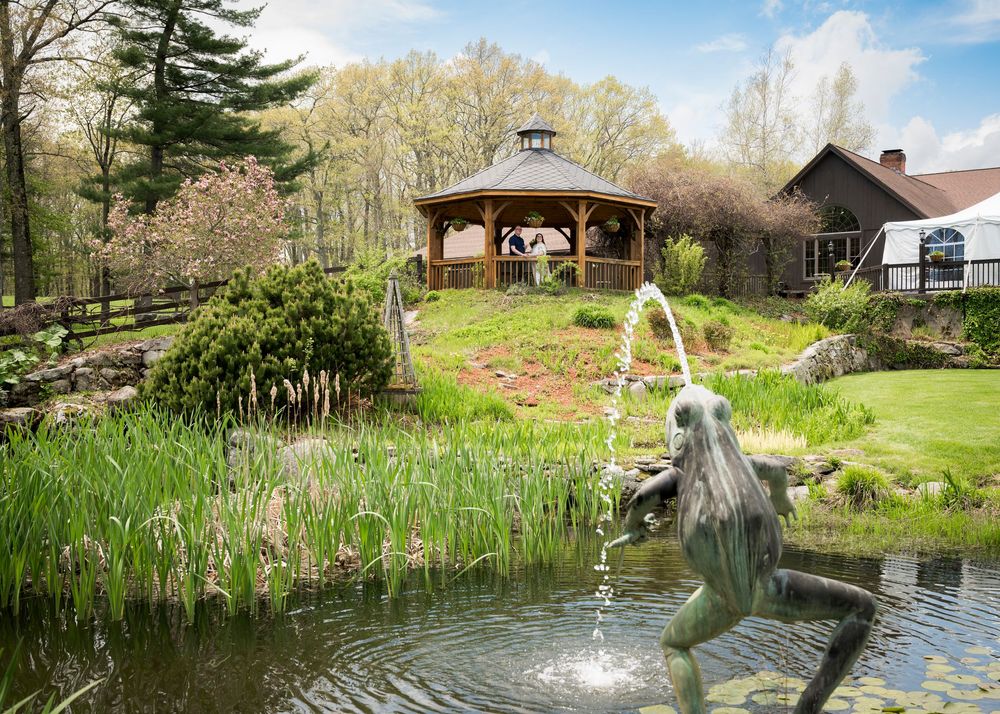 Scenic wedding venue with pond, dancing frog fountain, gazebo, and rustic barn surrounded by trees.