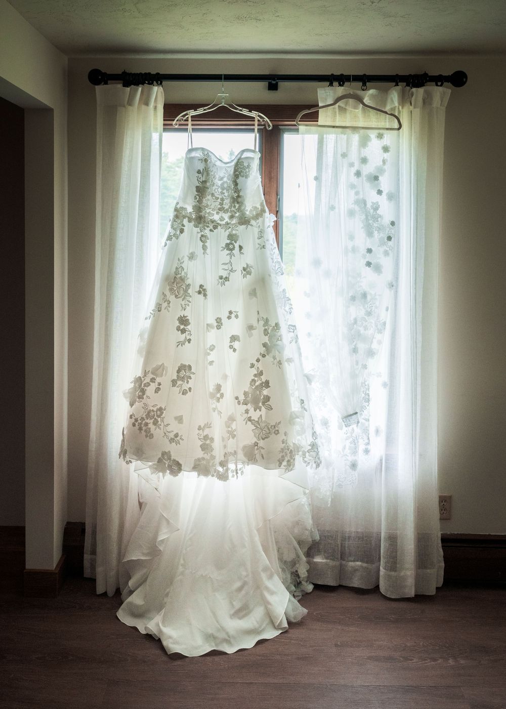 Strapless floral embroidered wedding gown hanging elegantly in front of sheer curtained window.