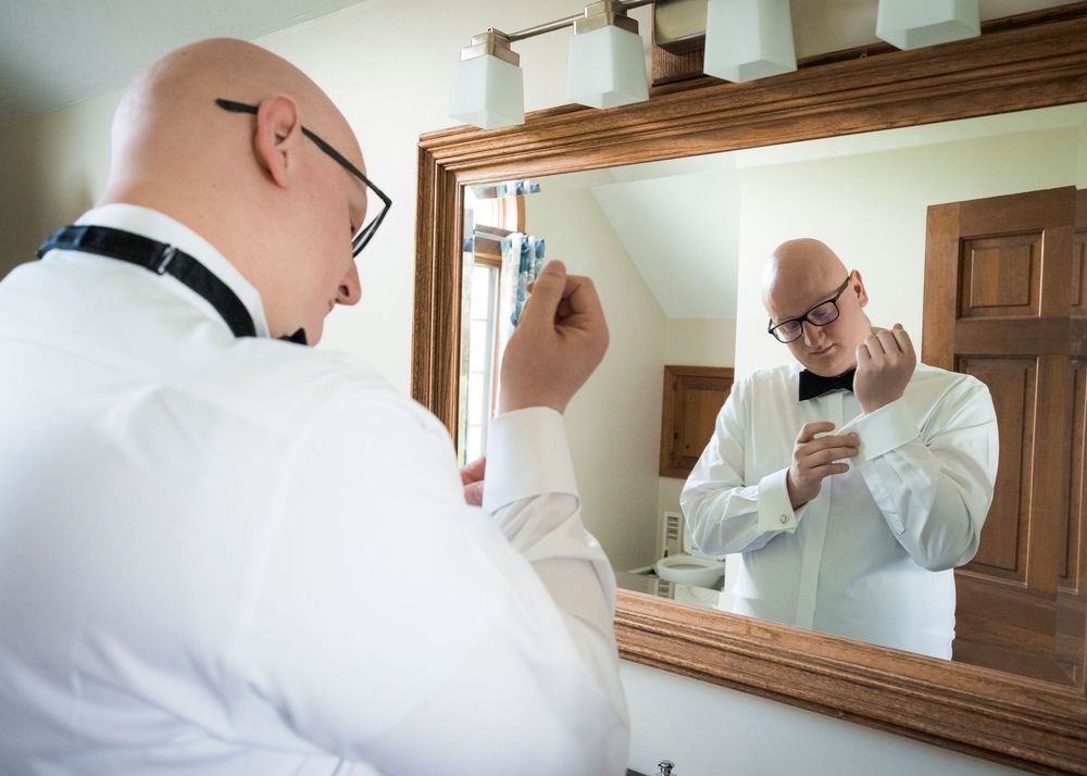 Bald groom in white shirt preparing in front of a mirror on wedding day.