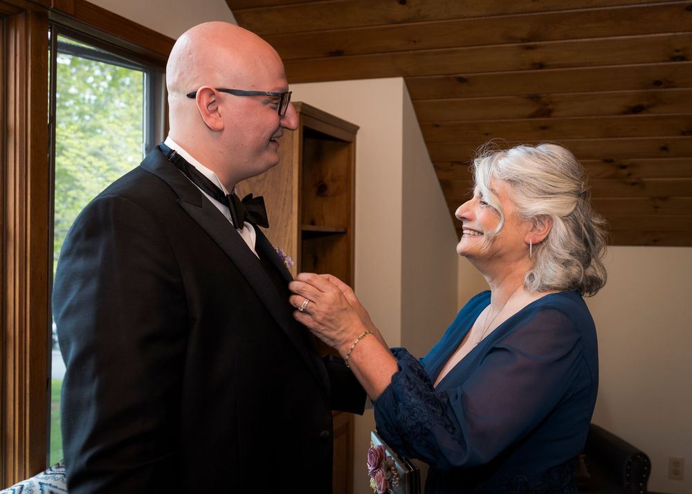 Older woman in navy dress adjusts grooms bow tie before wedding ceremony in rustic wood interior.