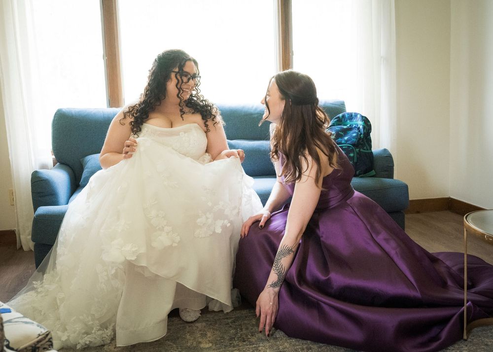 Bride in white gown chatting with bridesmaid in purple dress on a blue sofa.