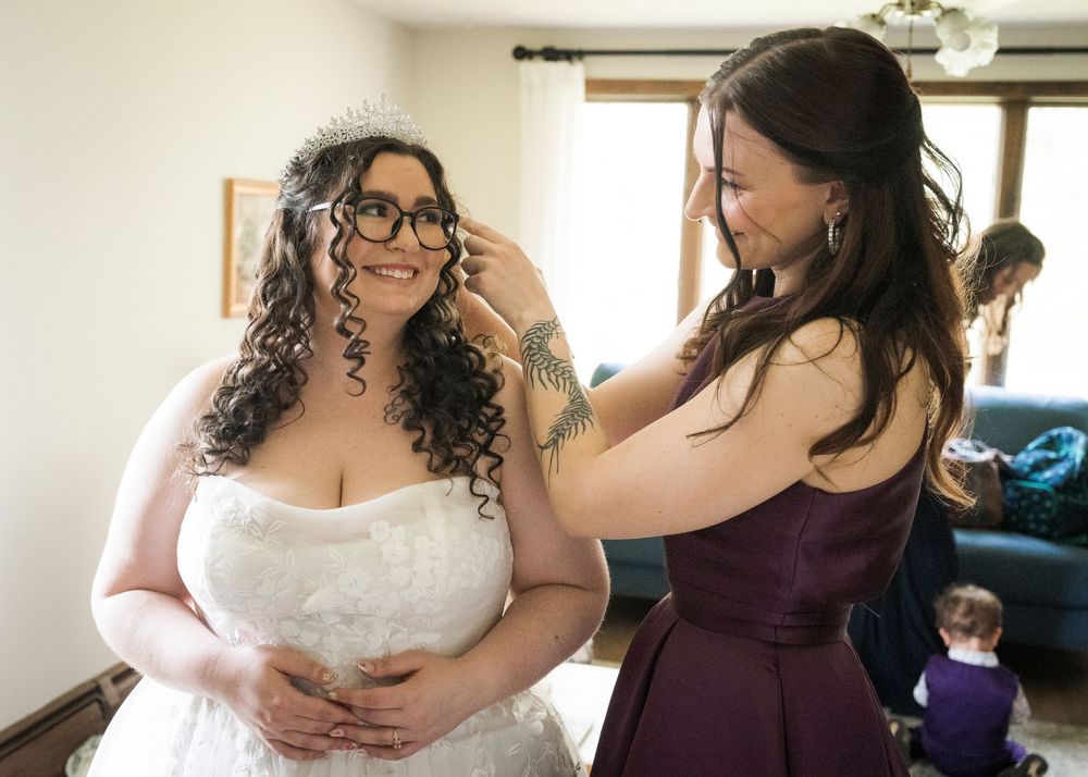 Bridesmaid helping bride adjust tiara while bride smiles in white lace wedding gown indoors.