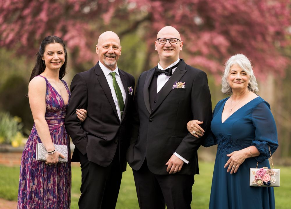 Two couples posing together outdoors in formal attire with pink cherry blossoms behind them.