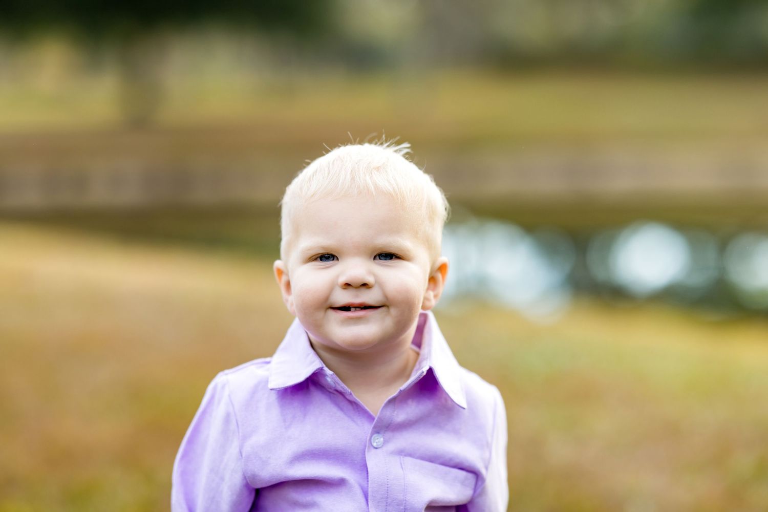 A baby in a purple collared shirt smiles while sitting outdoors in a natural setting.