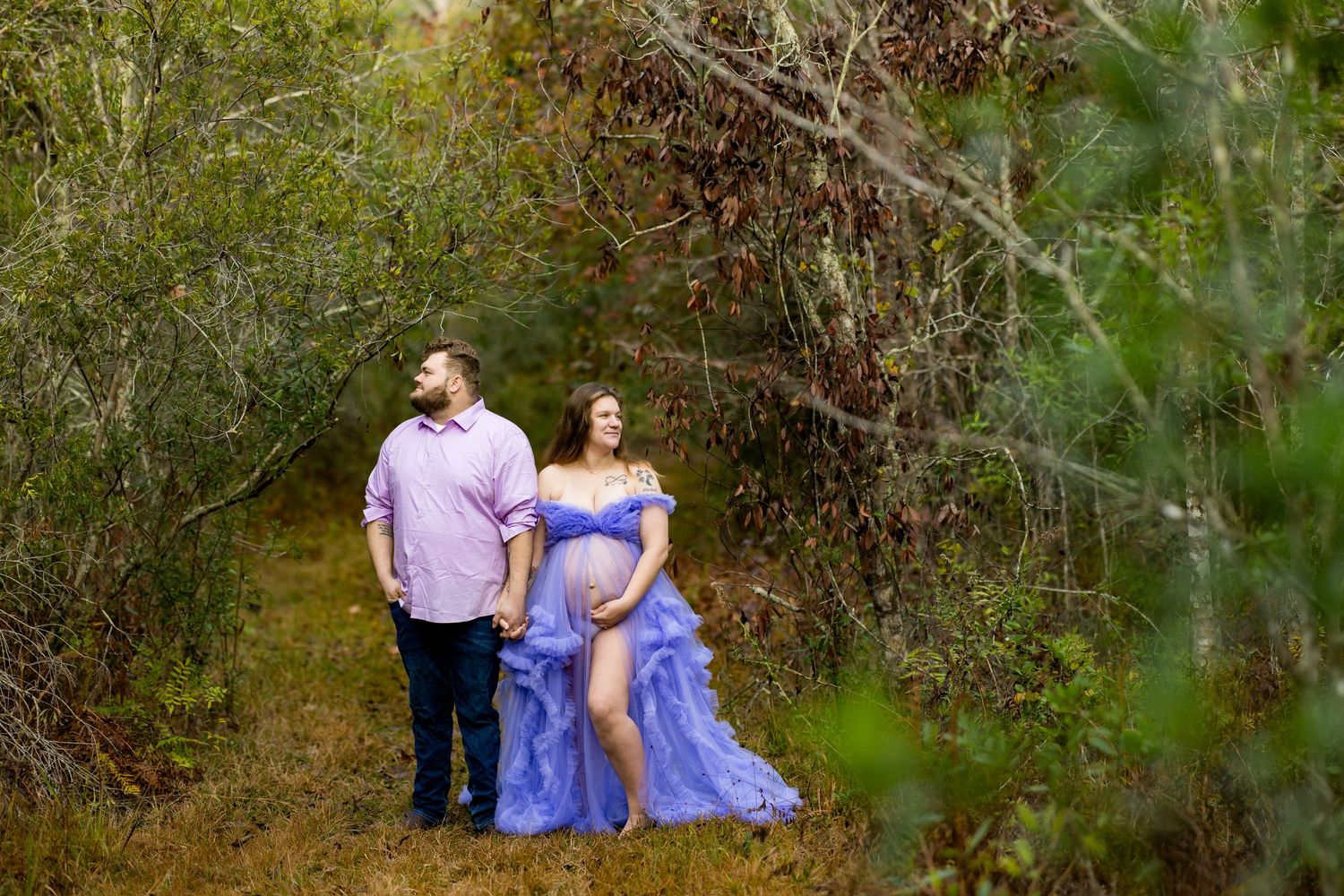 A couple in blue and pink outfits poses for romantic outdoor photos among green foliage in a natural setting.