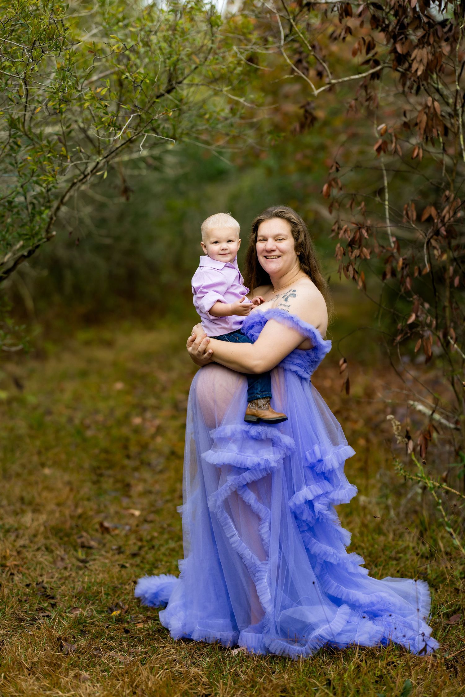 A woman in a flowing periwinkle blue tulle gown stands outdoors among autumn foliage with a baby.