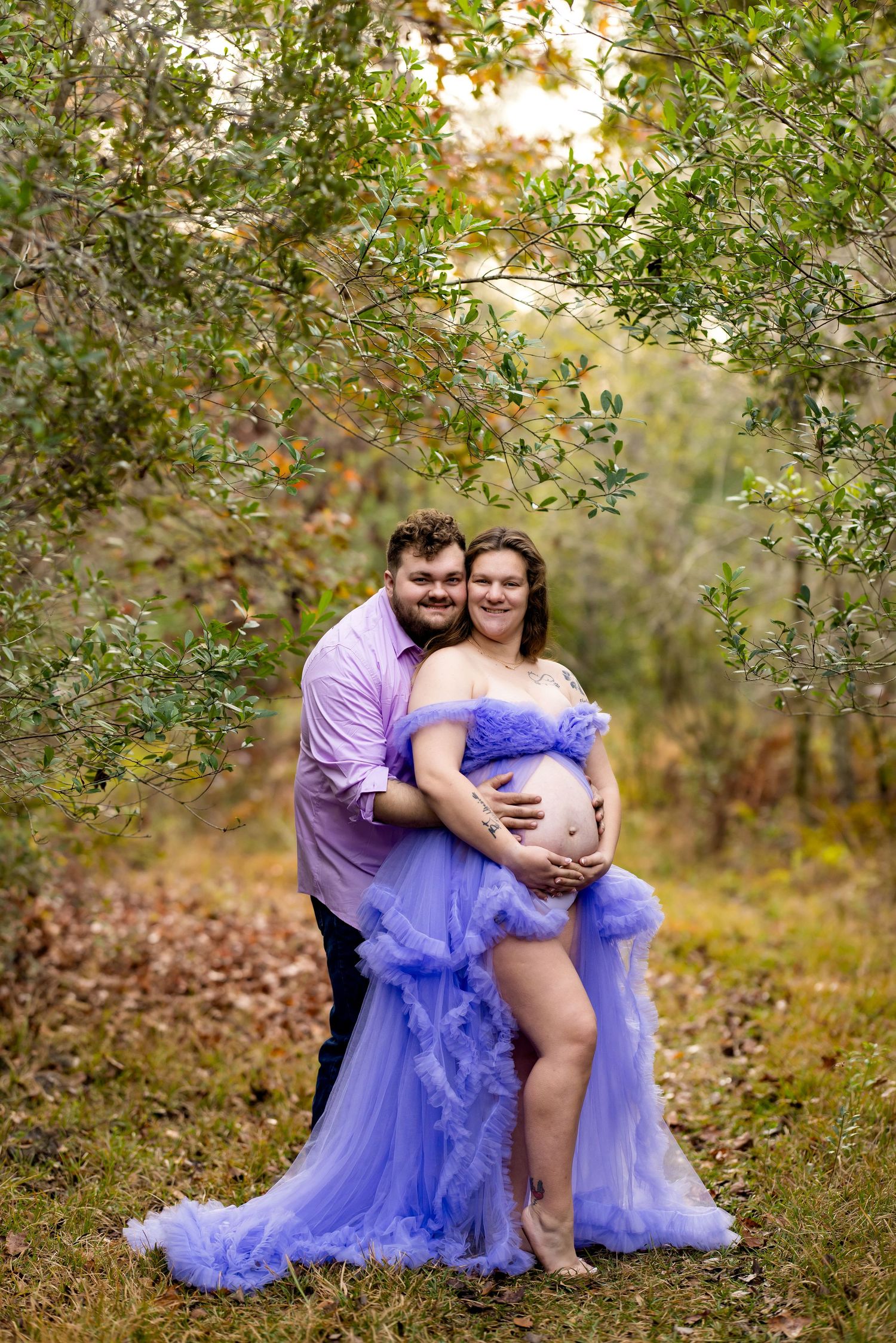 A couple in a purple dress and shirt embrace outdoors among autumn trees and fallen leaves.