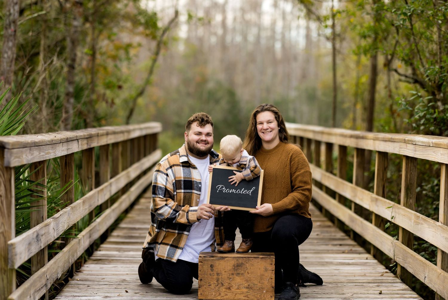 Two people kneel on a wooden bridge in a forest setting while holding a chalkboard sign.