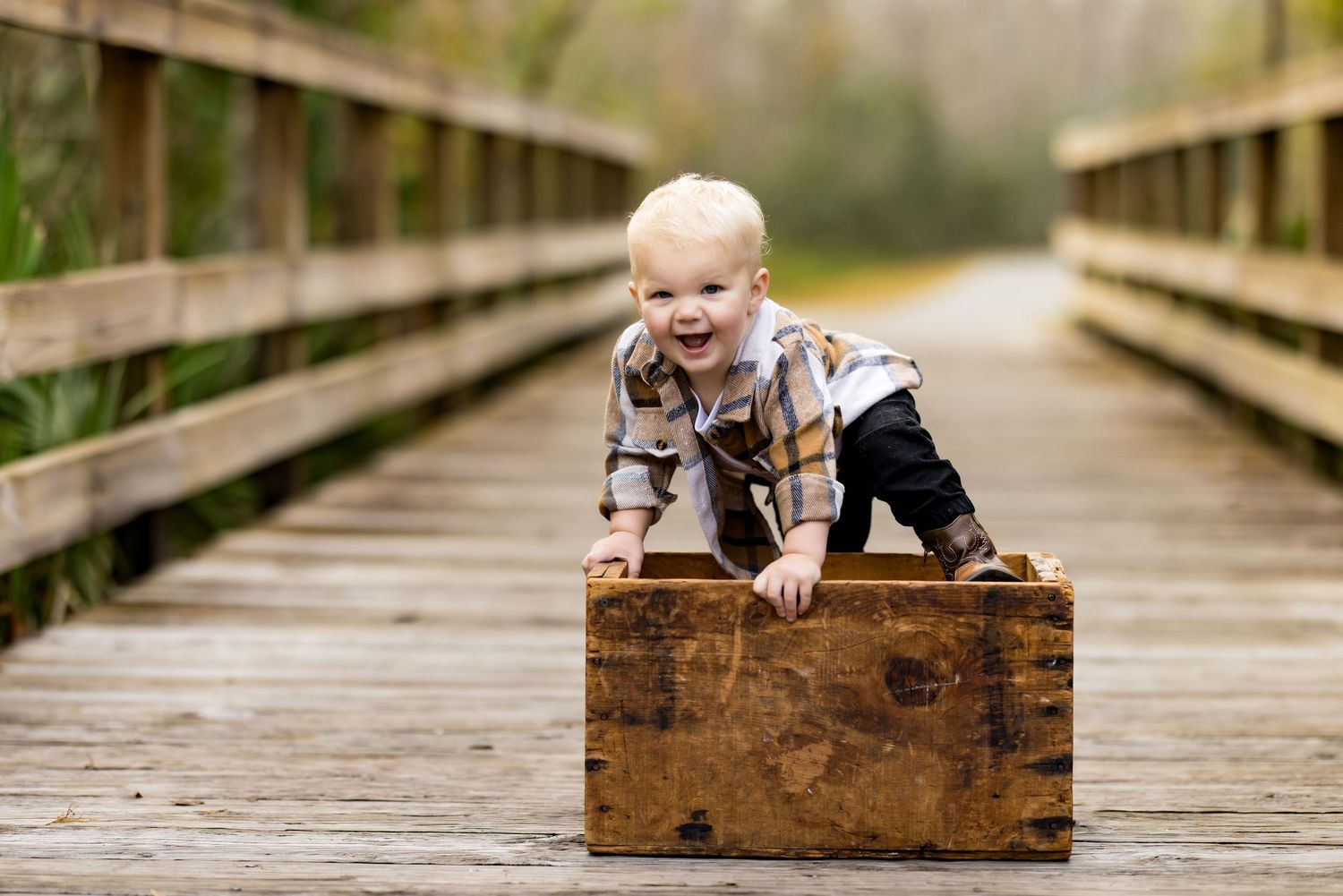 Baby in plaid shirt crawls on vintage wooden crate while exploring wooden bridge walkway outdoors.