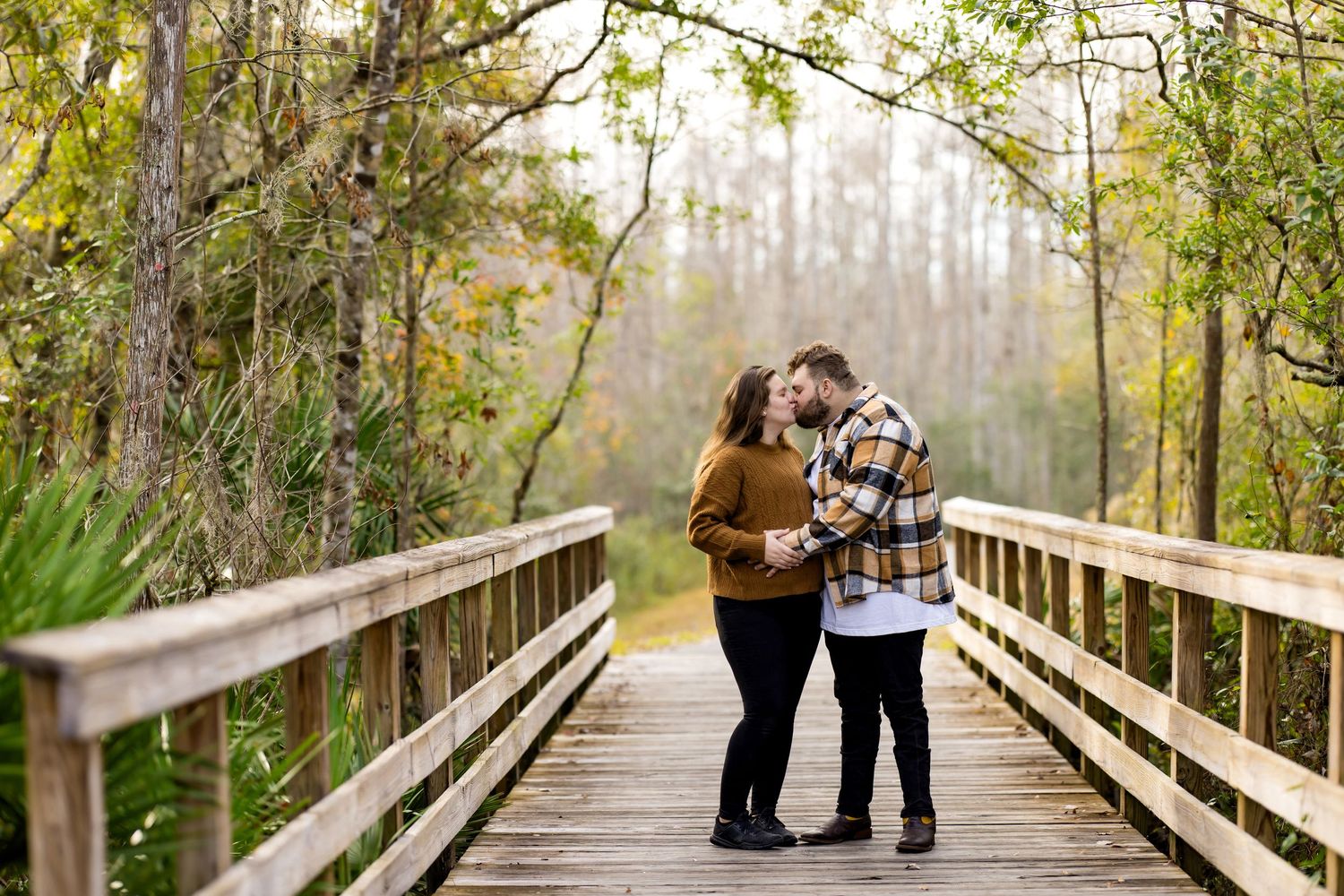 A couple stands close together on a wooden boardwalk surrounded by lush green forest on a misty day.