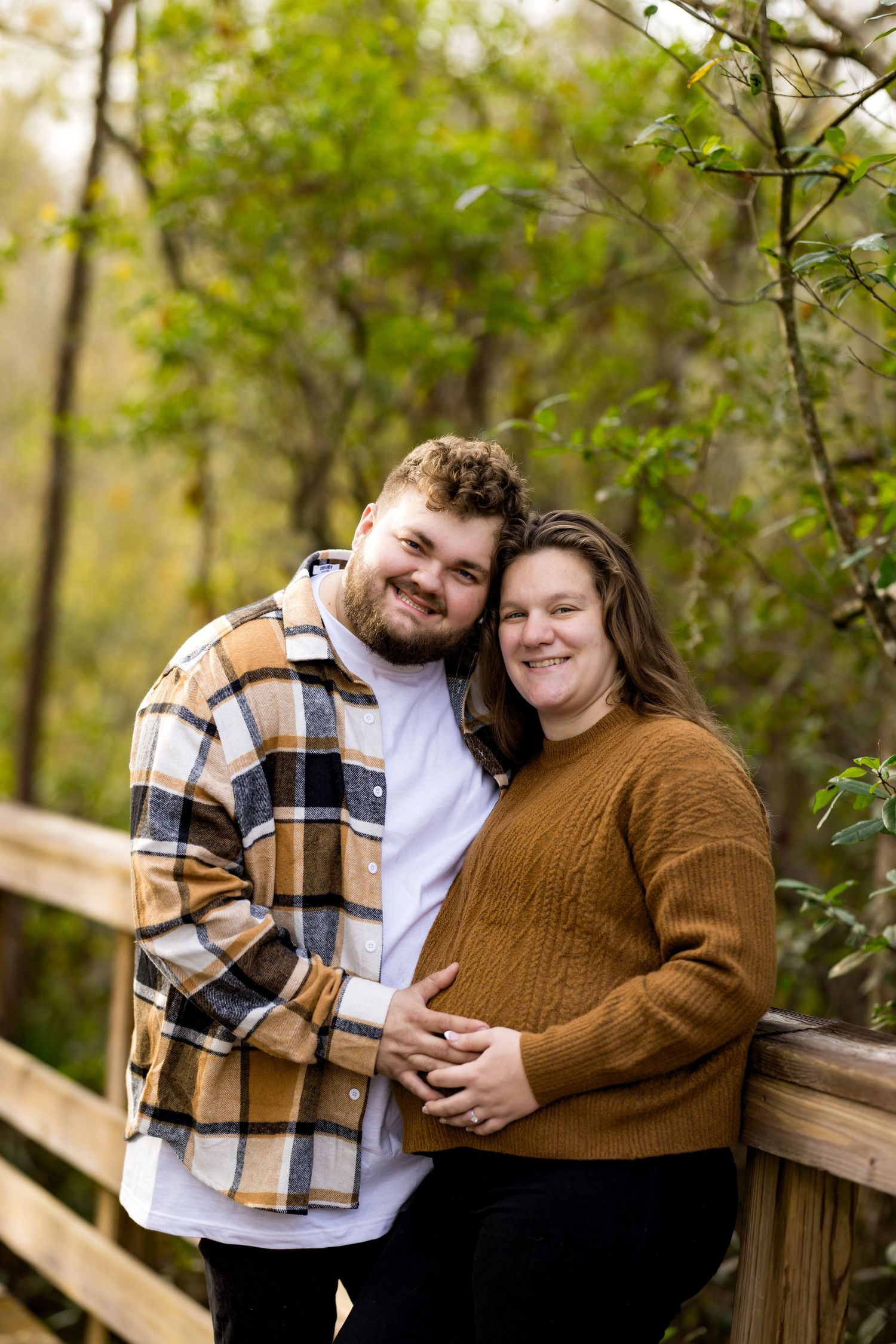 A couple in autumn attire embraces on a wooden bridge surrounded by green foliage in a natural setting.