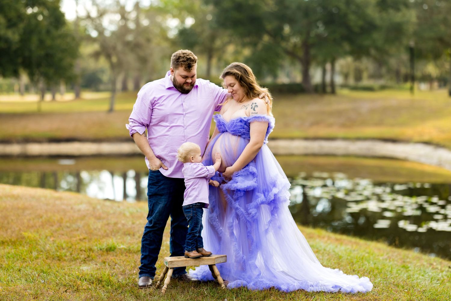 A couple in wedding attire poses together in a romantic outdoor setting with a lake and trees in the background.