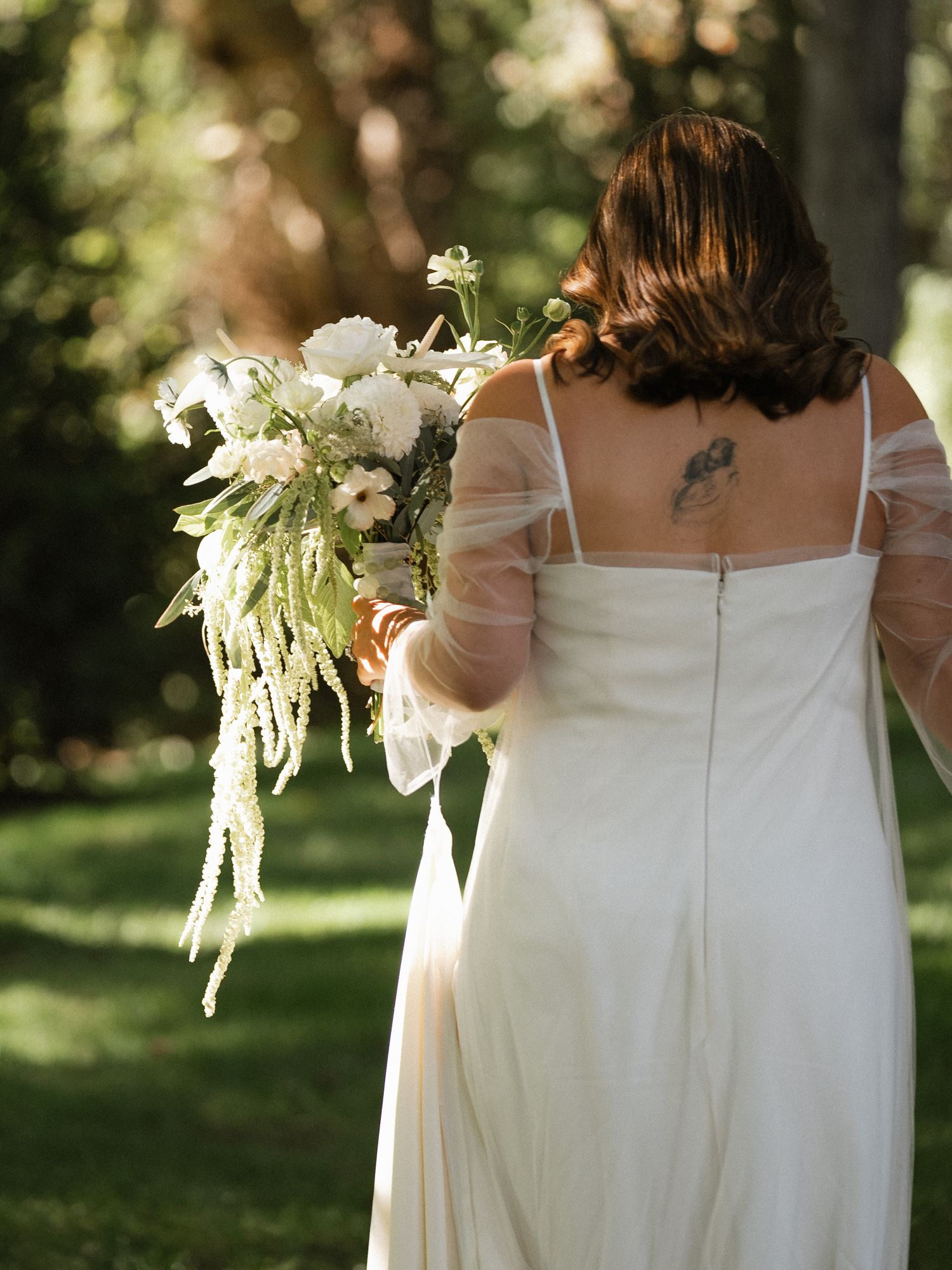 Back view of white flowing dresses with delicate floral bouquets against a sunlit garden backdrop.