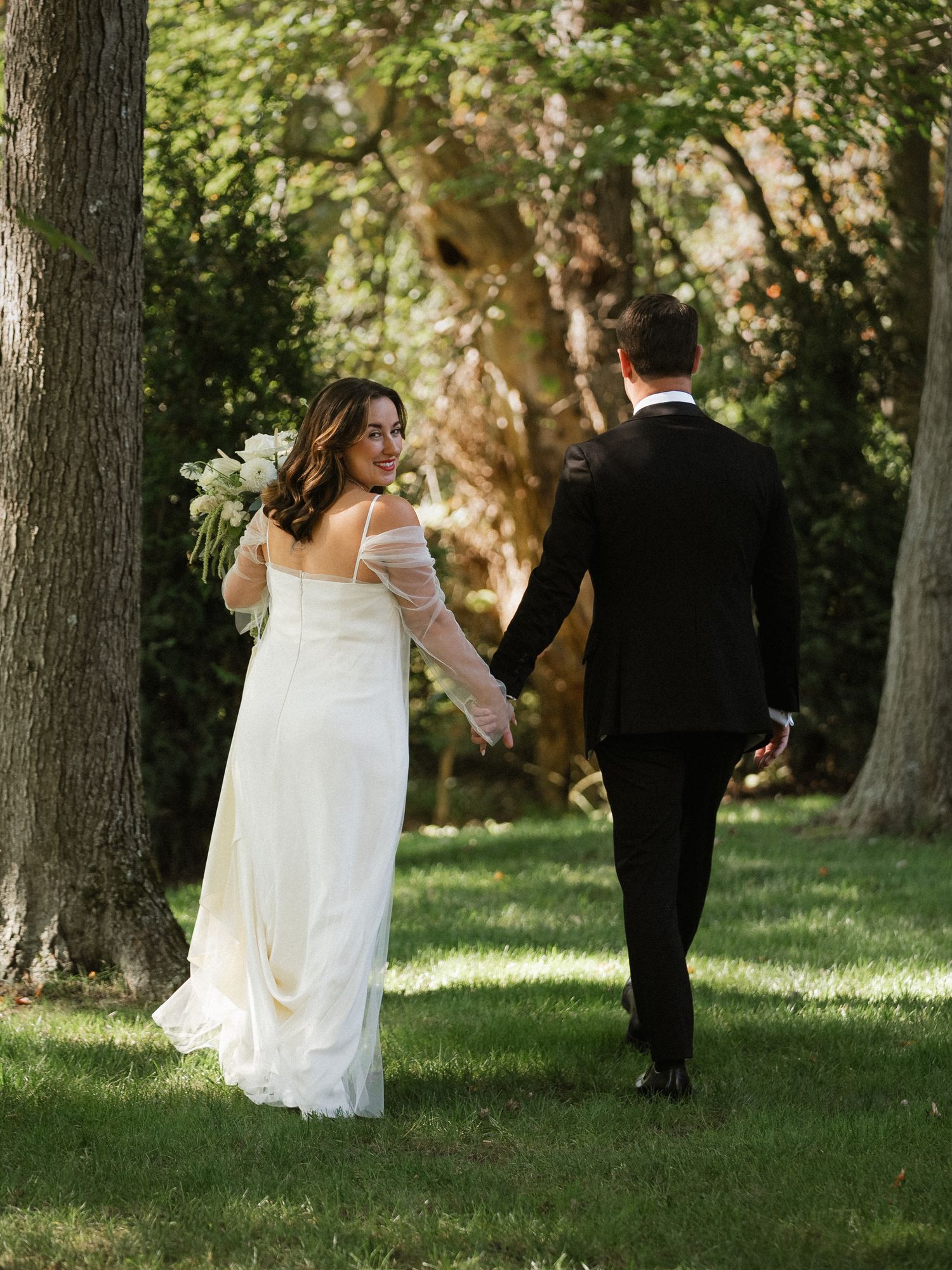 A romantic wedding couple dances together on a sunlit lawn surrounded by tall trees.