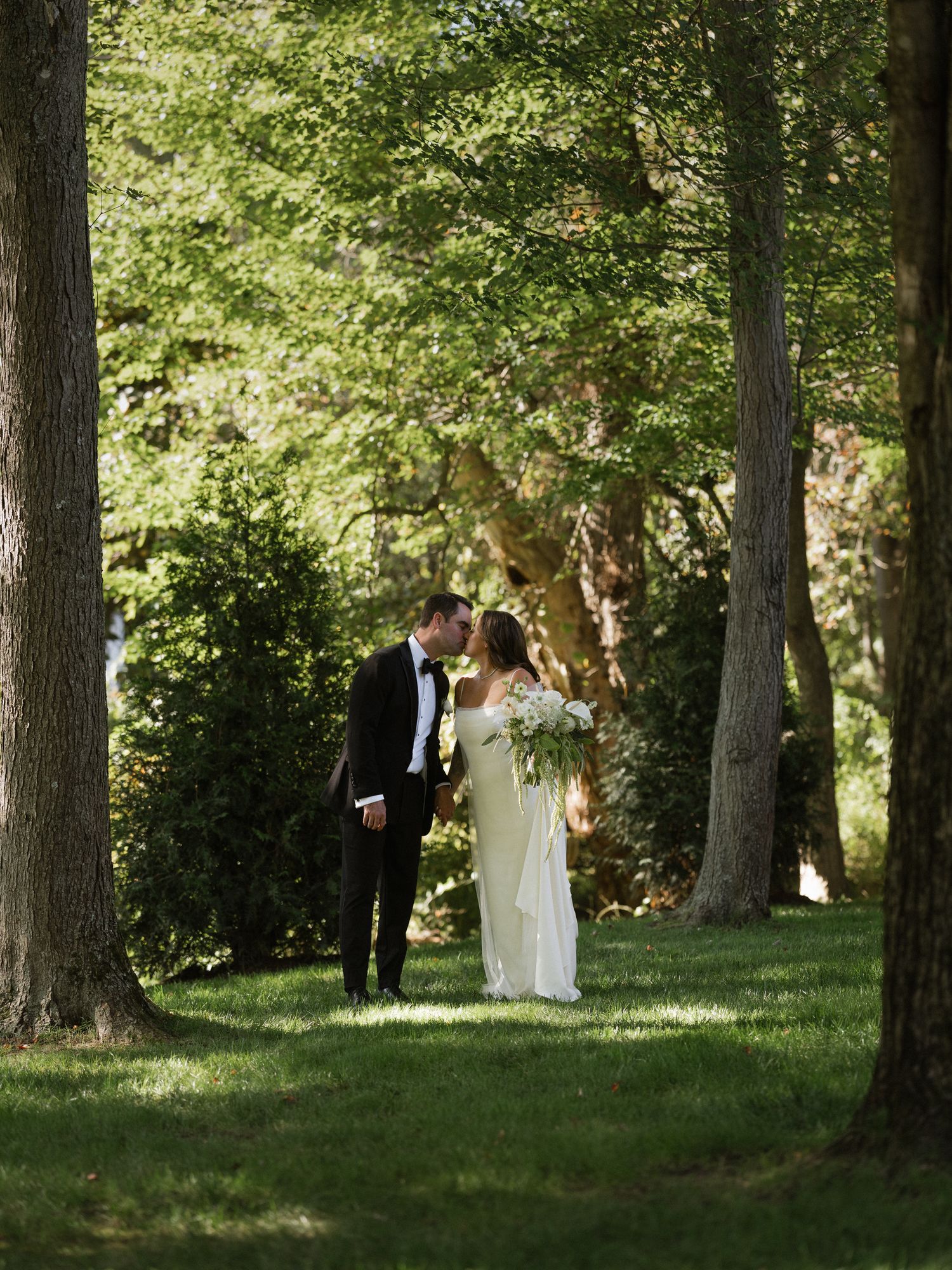 Wedding couple walks through a sunlit wooded area with tall trees creating a natural outdoor setting.