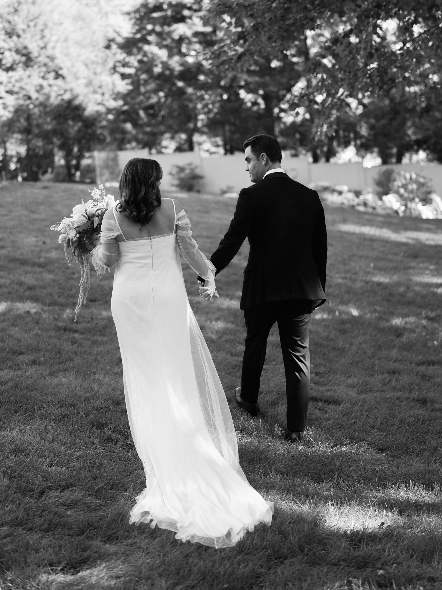 Wedding couple walking through a grassy field with the bride's veil flowing in black and white photography.