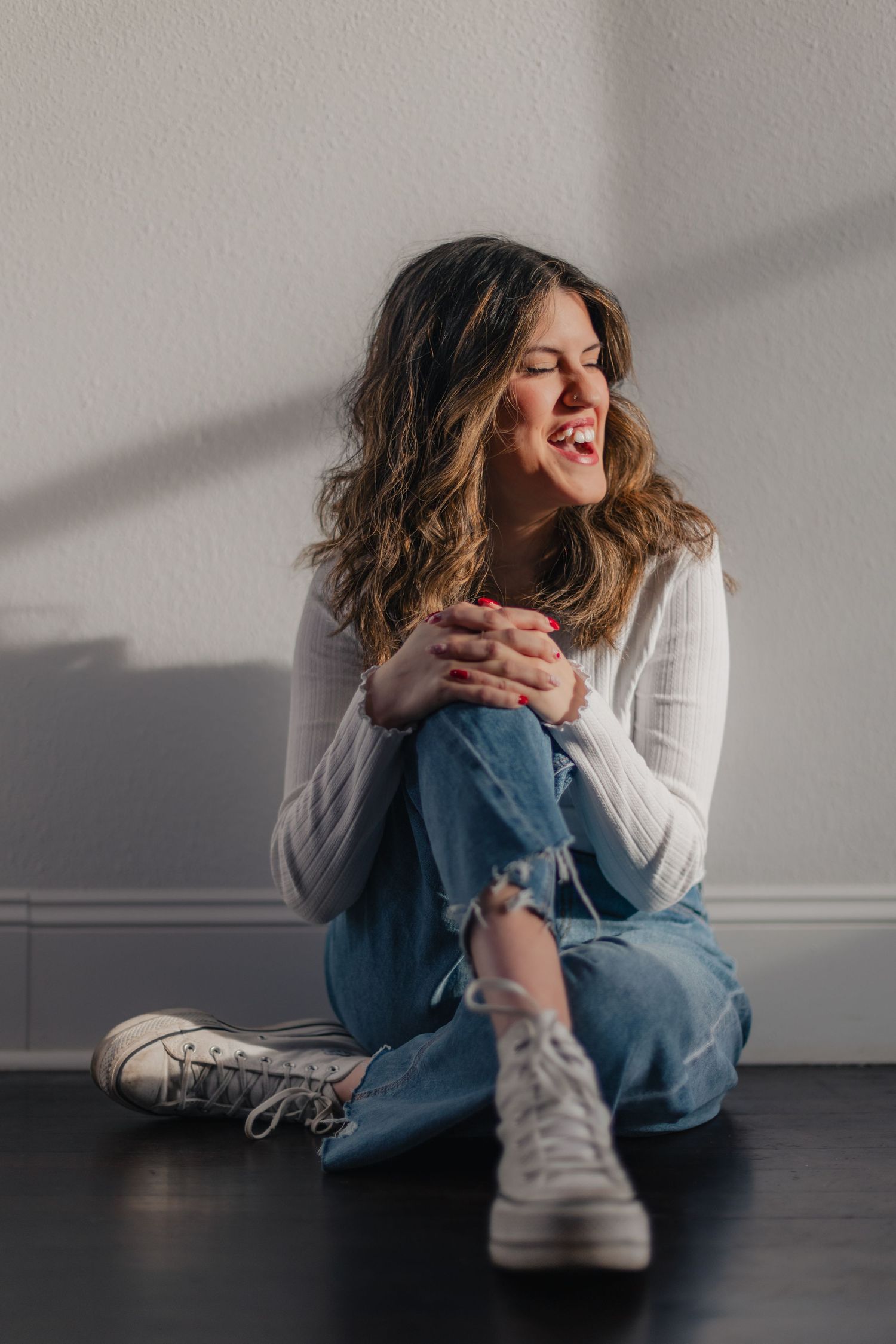 A person in a white sweater and jeans laughs while sitting on a dark floor in a minimalist setting.