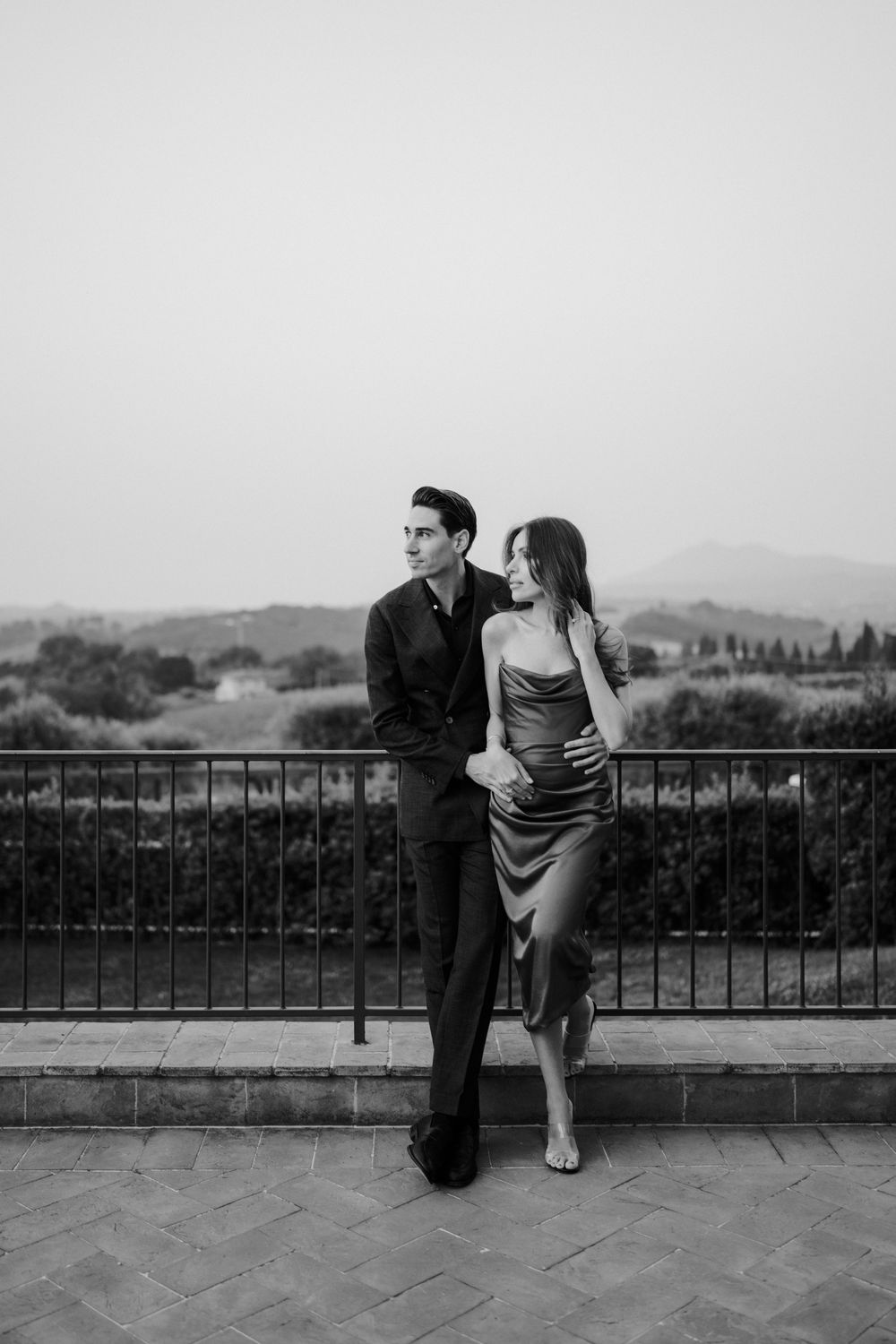 A romantic couple shares a tender moment on a balcony overlooking rolling hills in black and white.