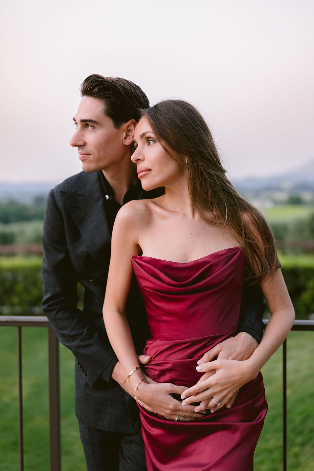 A romantic couple poses together outdoors, with the woman wearing a burgundy silk dress against a scenic landscape background.