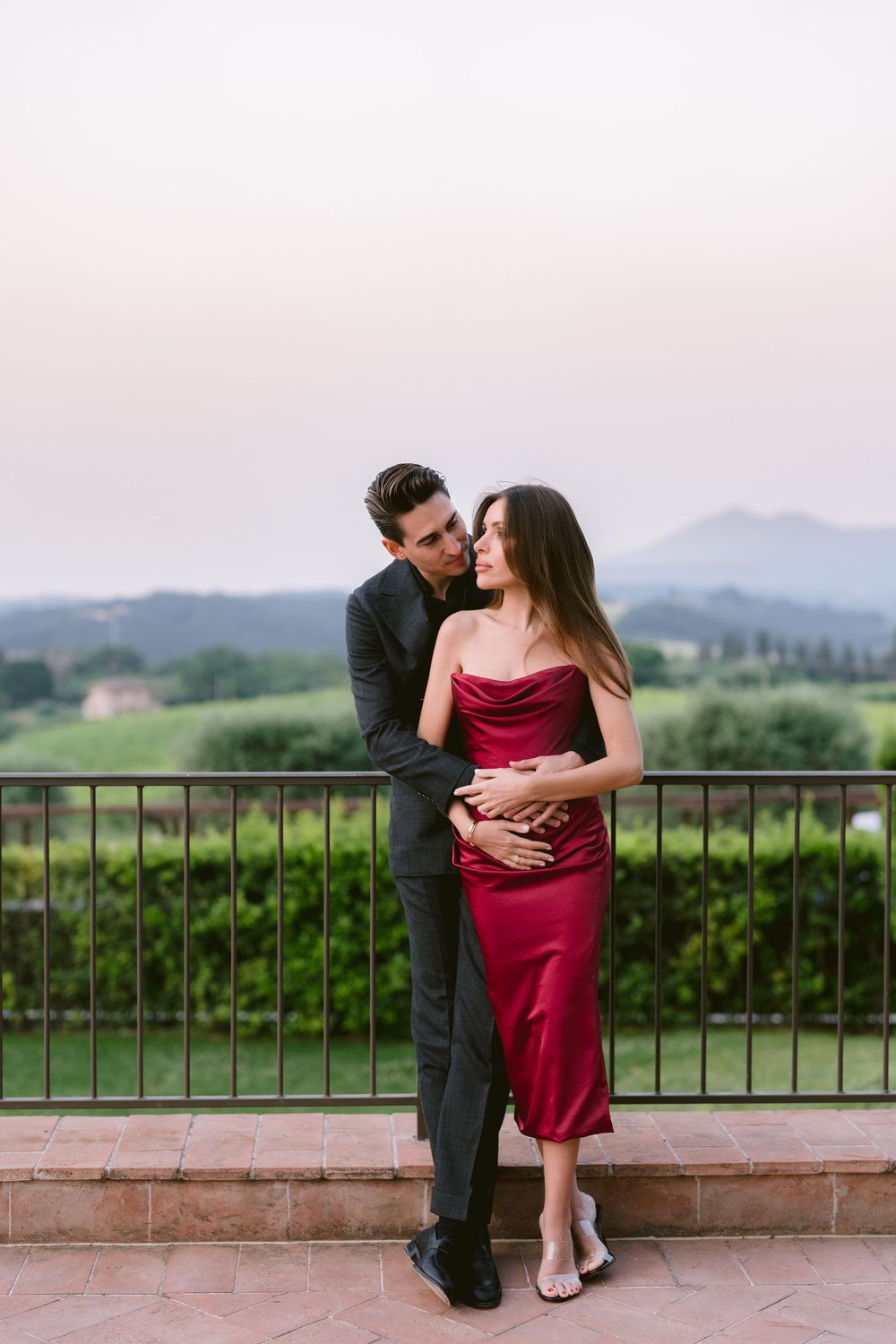 Romantic couple in formal attire embraces against scenic mountain backdrop at sunset on outdoor terrace.