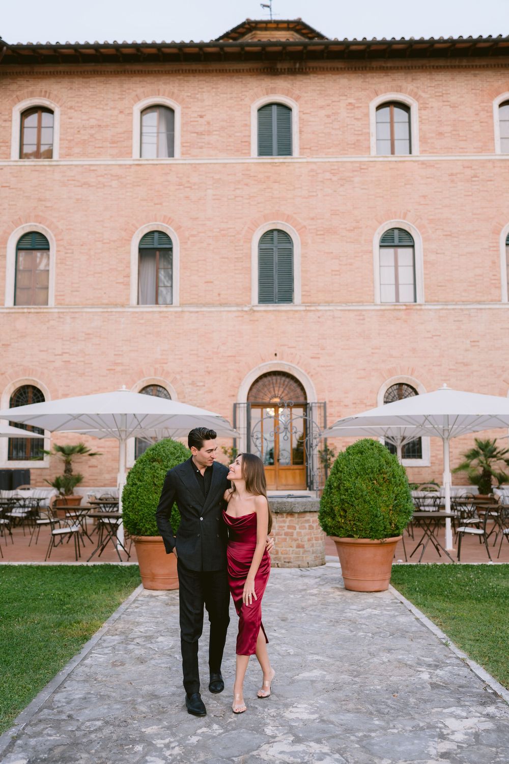 A couple walks together on a stone path outside a pink Italian villa with manicured gardens and arched windows.