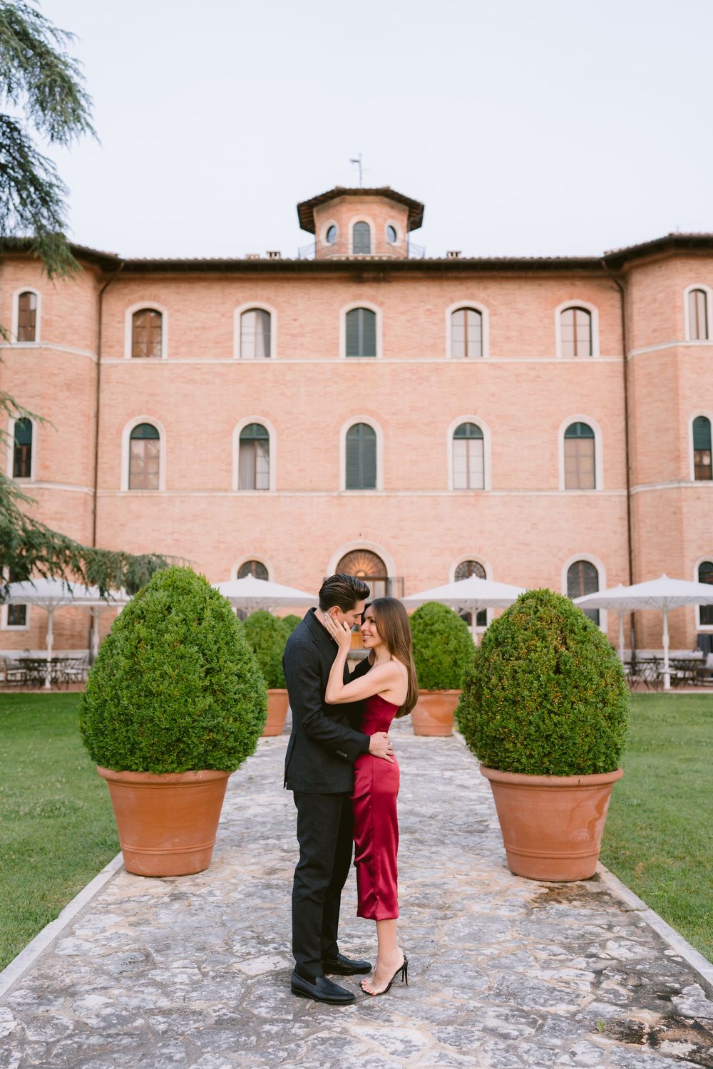A couple shares a romantic moment on a stone path in front of a Mediterranean-style villa with manicured topiary bushes.