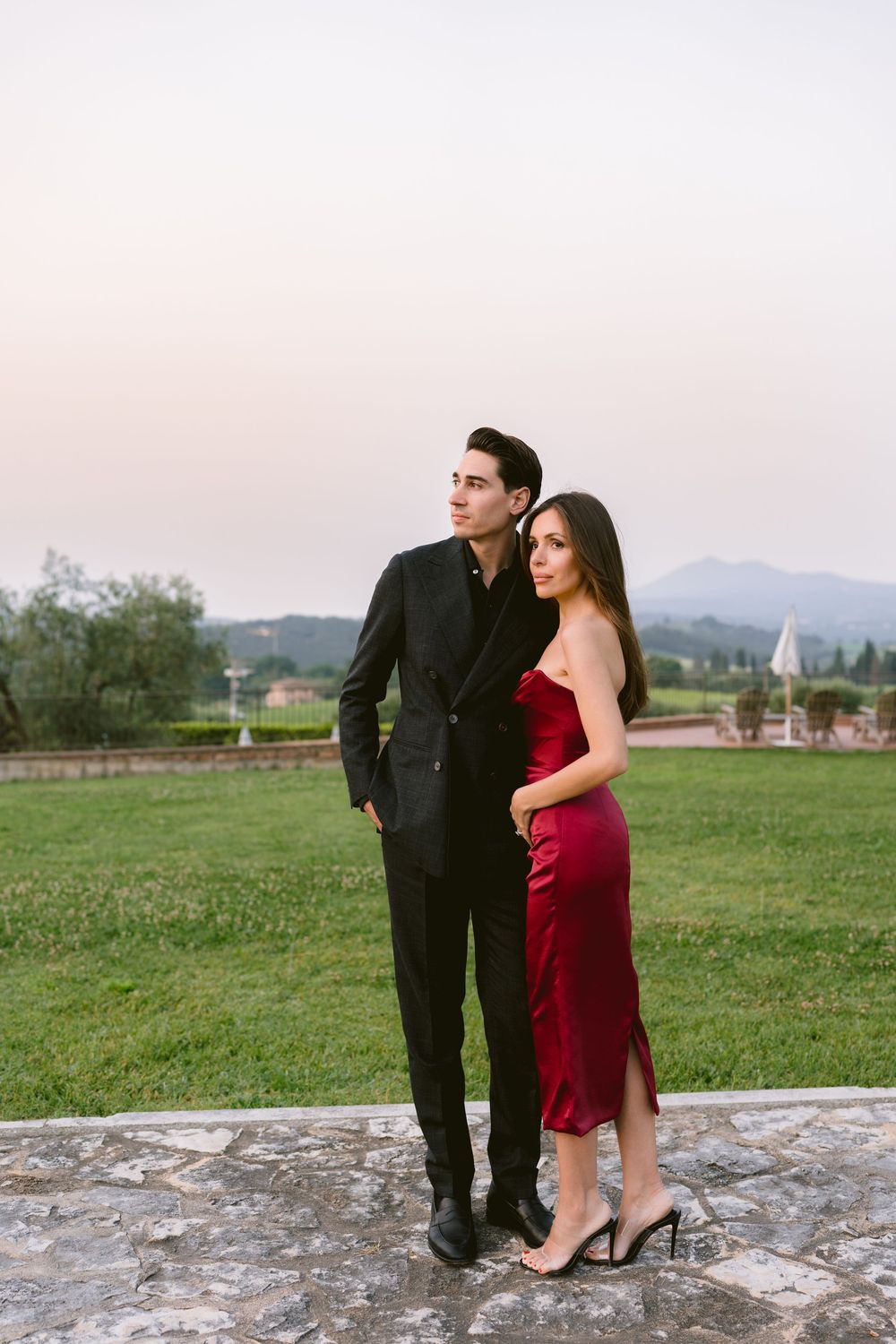 A couple in formal attire poses together on a stone surface with rolling hills and mountains in the background at sunset.