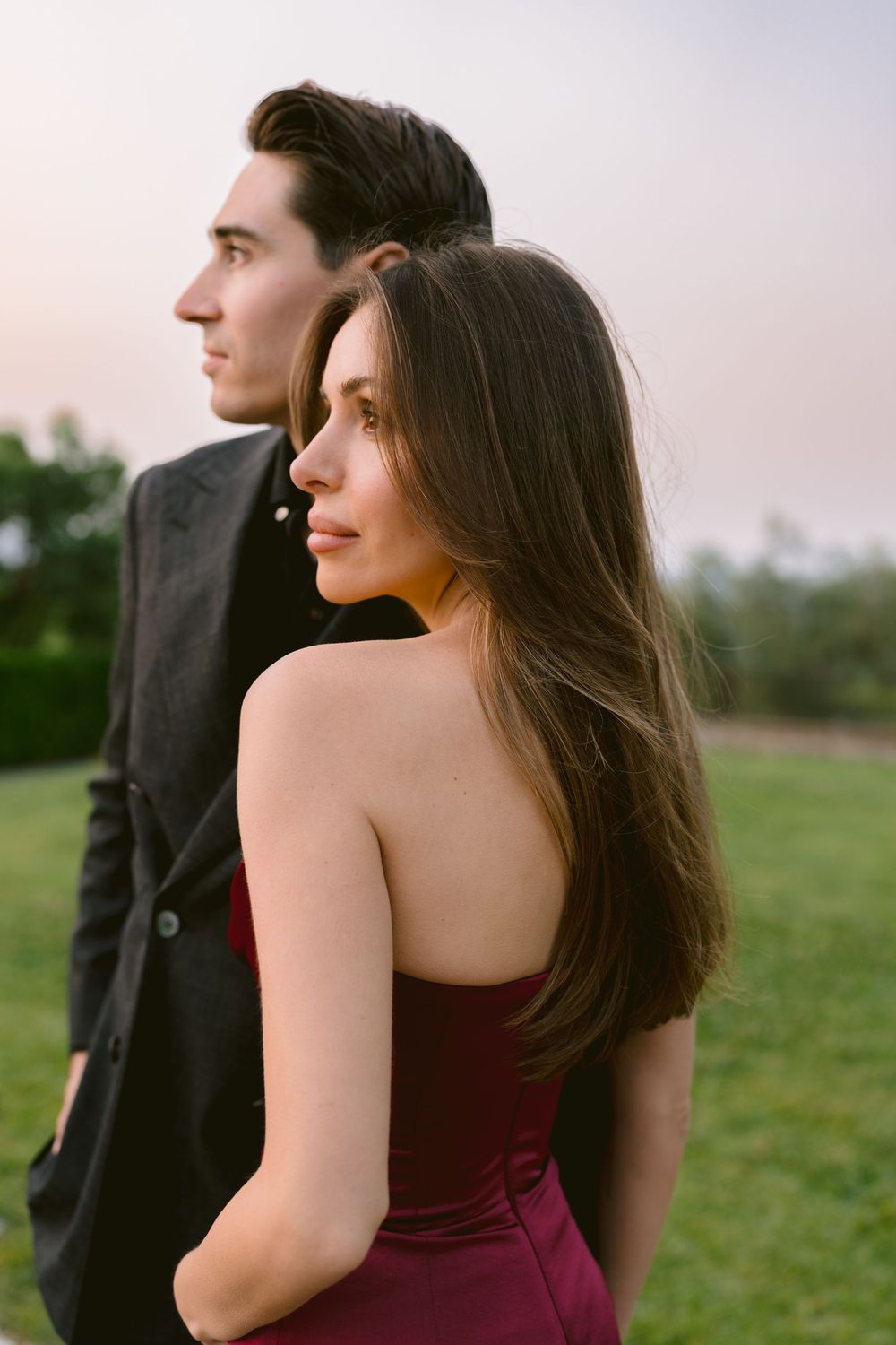A couple in formal attire poses in a grassy field at sunset with the person in burgundy looking away from the camera.