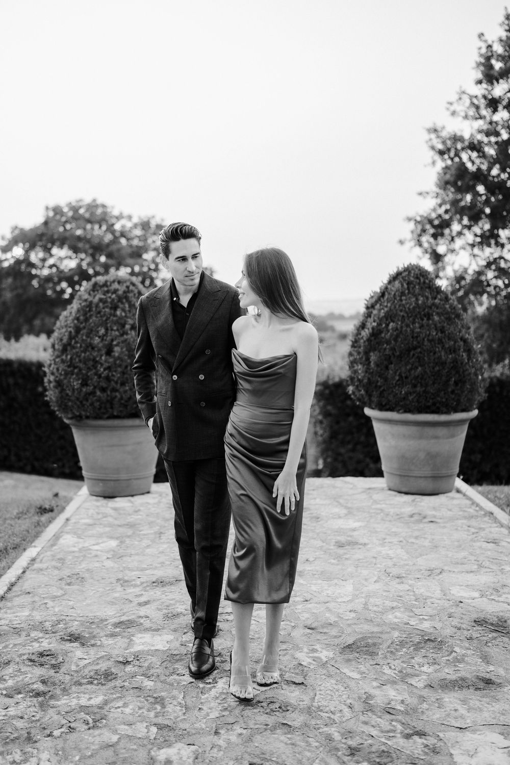 A couple stands together in a garden setting with decorative potted shrubs in black and white.