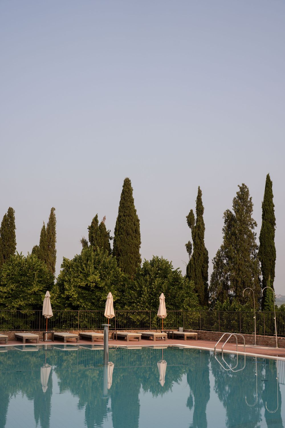 Tranquil swimming pool surrounded by tall cypress trees and lounge chairs with beige umbrellas against a serene sky.