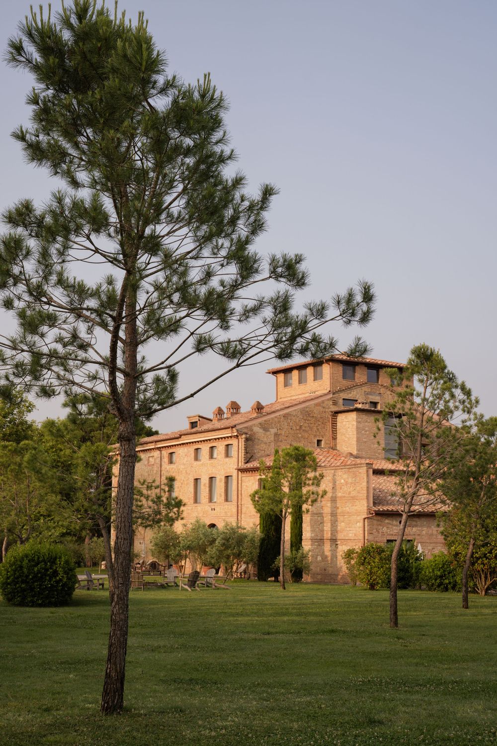 A tall pine tree stands in front of a historic Mediterranean-style villa with terracotta walls and tower in golden sunlight.