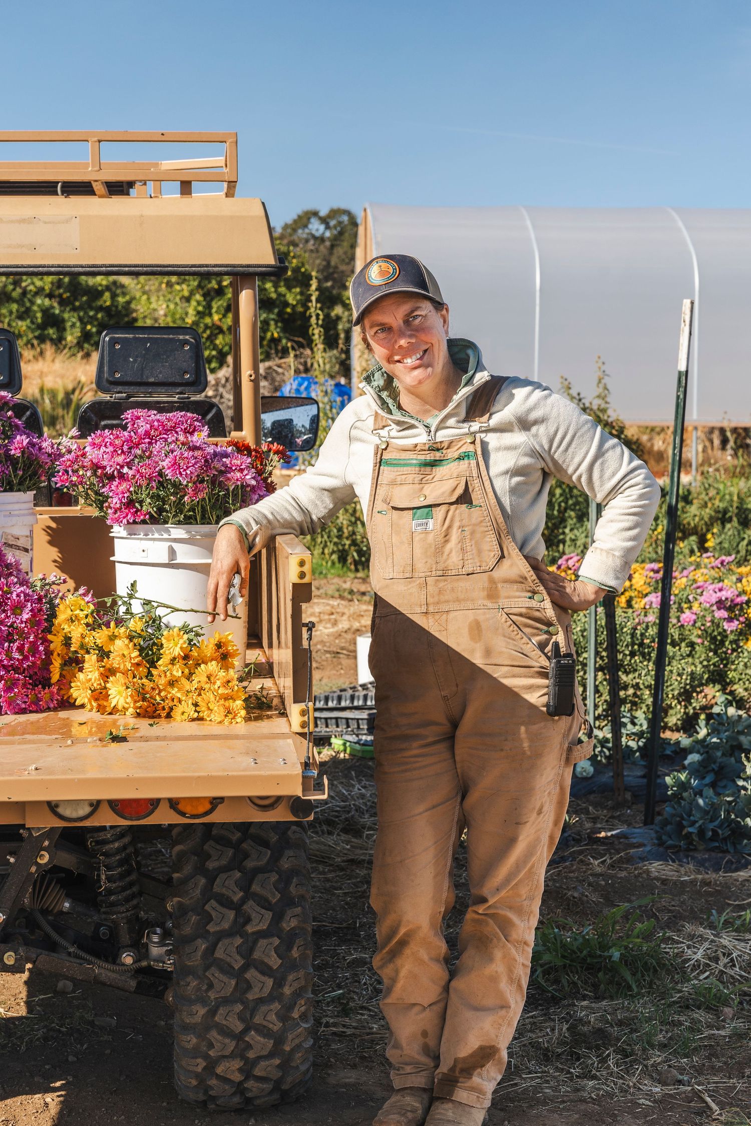 A person in brown overalls poses playfully near a rustic wooden cart in a colorful flower garden.