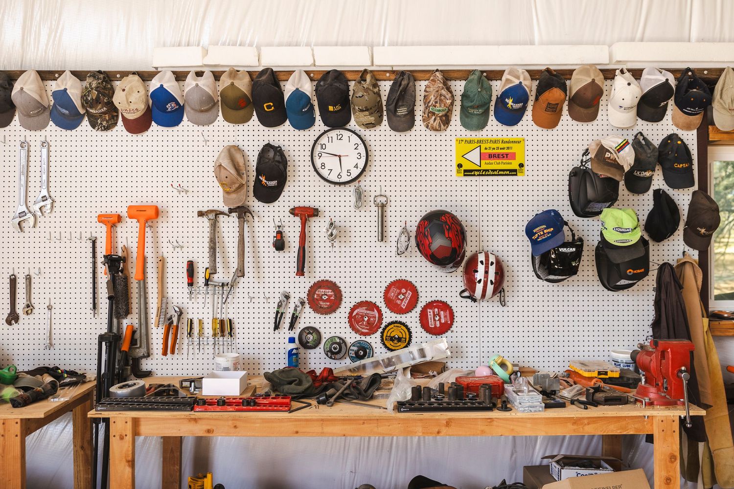 A workshop wall displays a large collection of baseball caps and hats mounted on pegboard with tools and equipment visible.