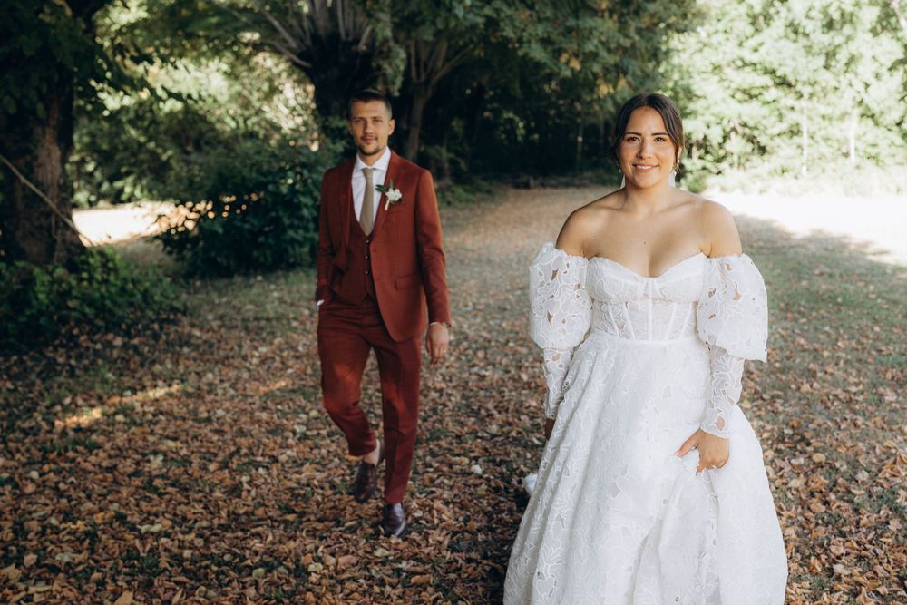 A romantic autumn wedding scene with a couple walking through fallen leaves in burgundy and white attire.