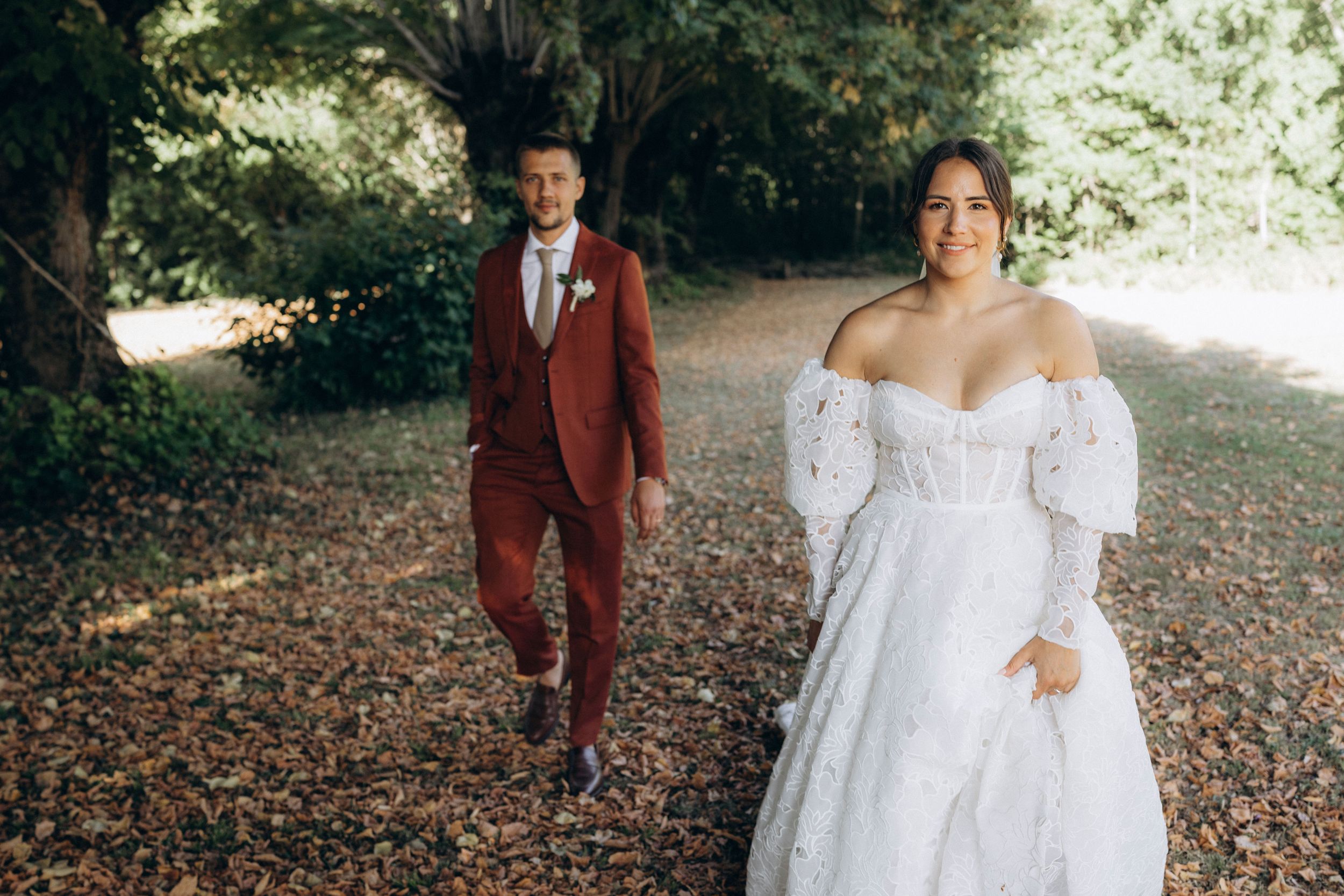 A romantic autumn wedding scene with a couple walking through fallen leaves in burgundy and white attire.