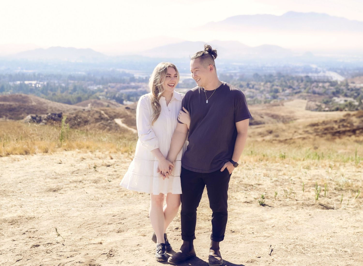 Couple posing on a scenic hillside with mountain views, woman in white dress and man in dark outfit.