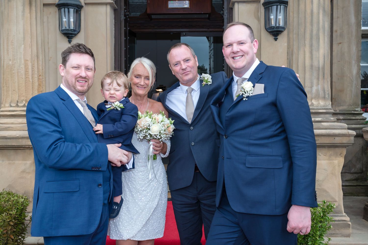 A group of wedding guests in formal attire gather outside DeVere Cranage Estate for a group photograph