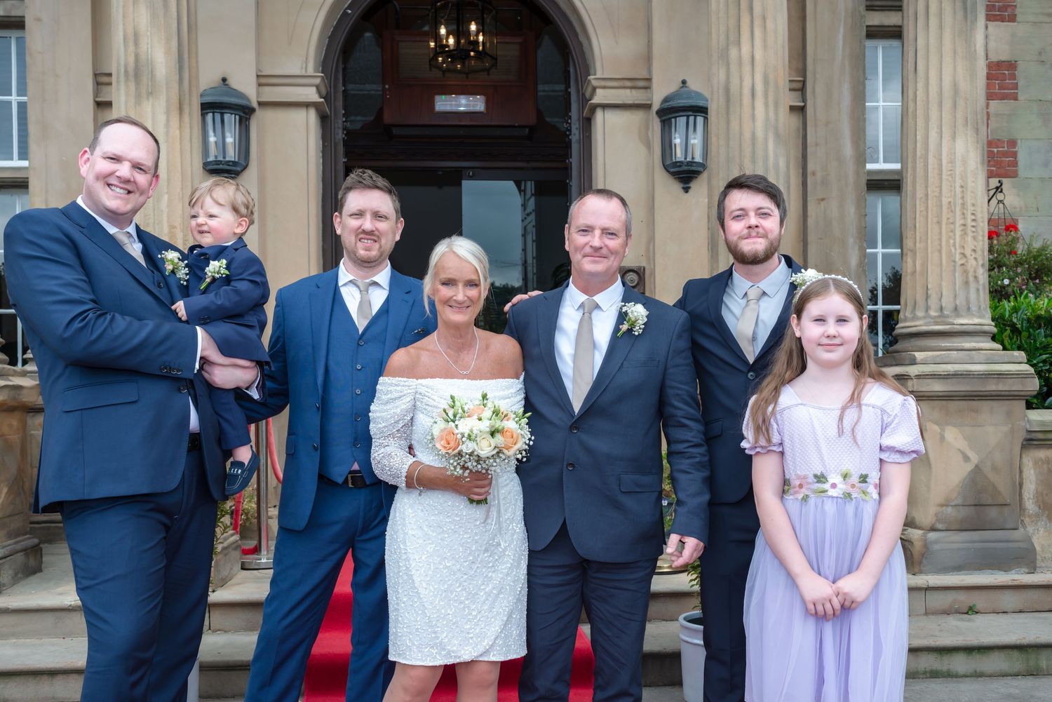 A group poses together in formal attire outside a grand stone building with ornate architectural details.