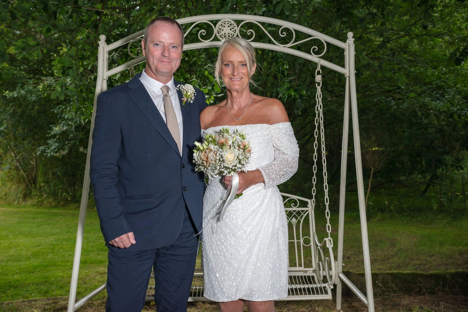Newlyweds posing under the white wrought iron wedding arch in the gardens at De Vere Cranage Estate; romantic Cheshire wedding photography