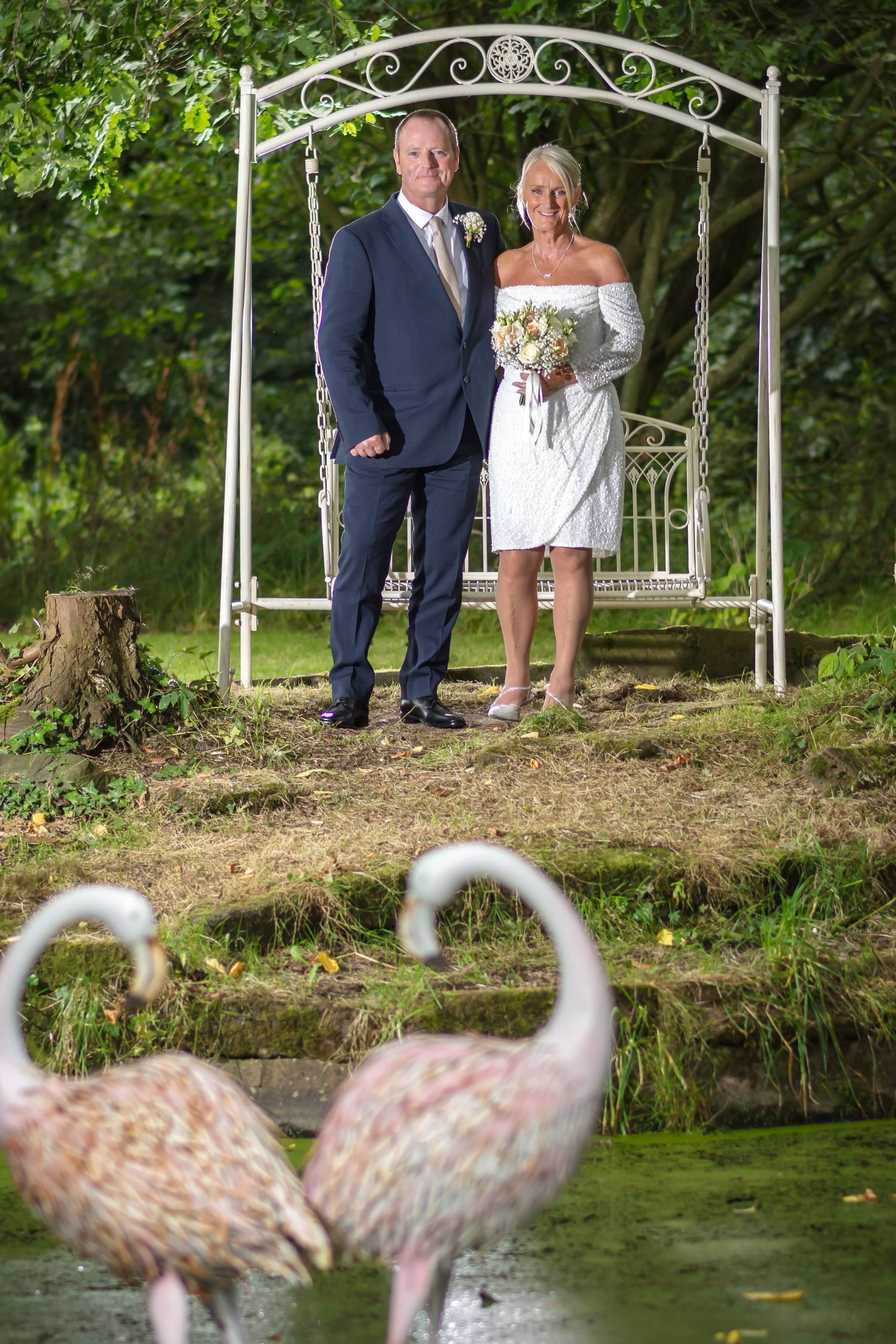 A couple poses near a pond with decorative white metal arbors and pink flamingos in a garden setting.