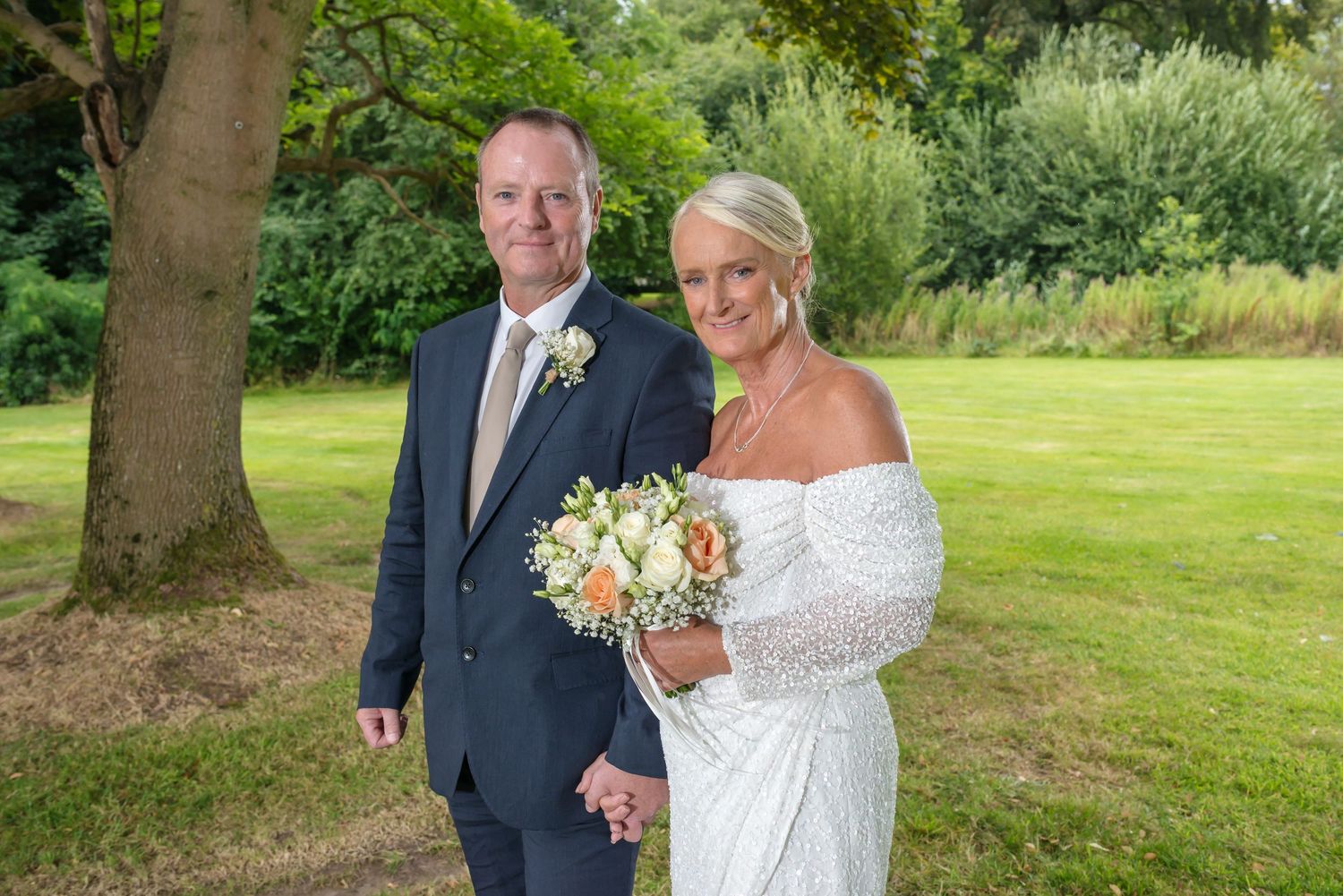 A couple in wedding attire poses together outdoors in a lush garden setting with tall trees in the background at DeVere Cranage Estate