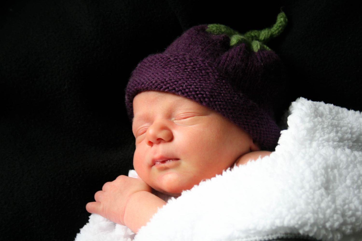 A sleeping newborn baby wrapped in white blankets against a black background.